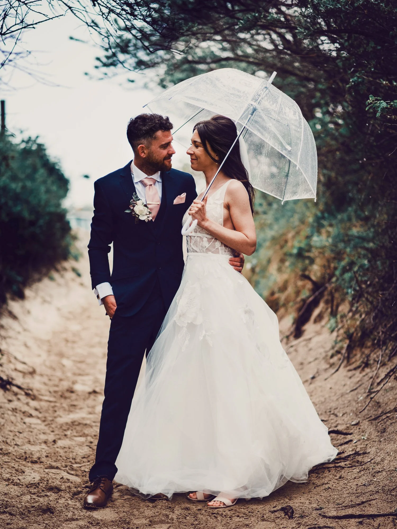 A bride in a white wedding gown and a groom in a dark suit under a transparent umbrella on a sandy path surrounded by greenery, on a rainy day.