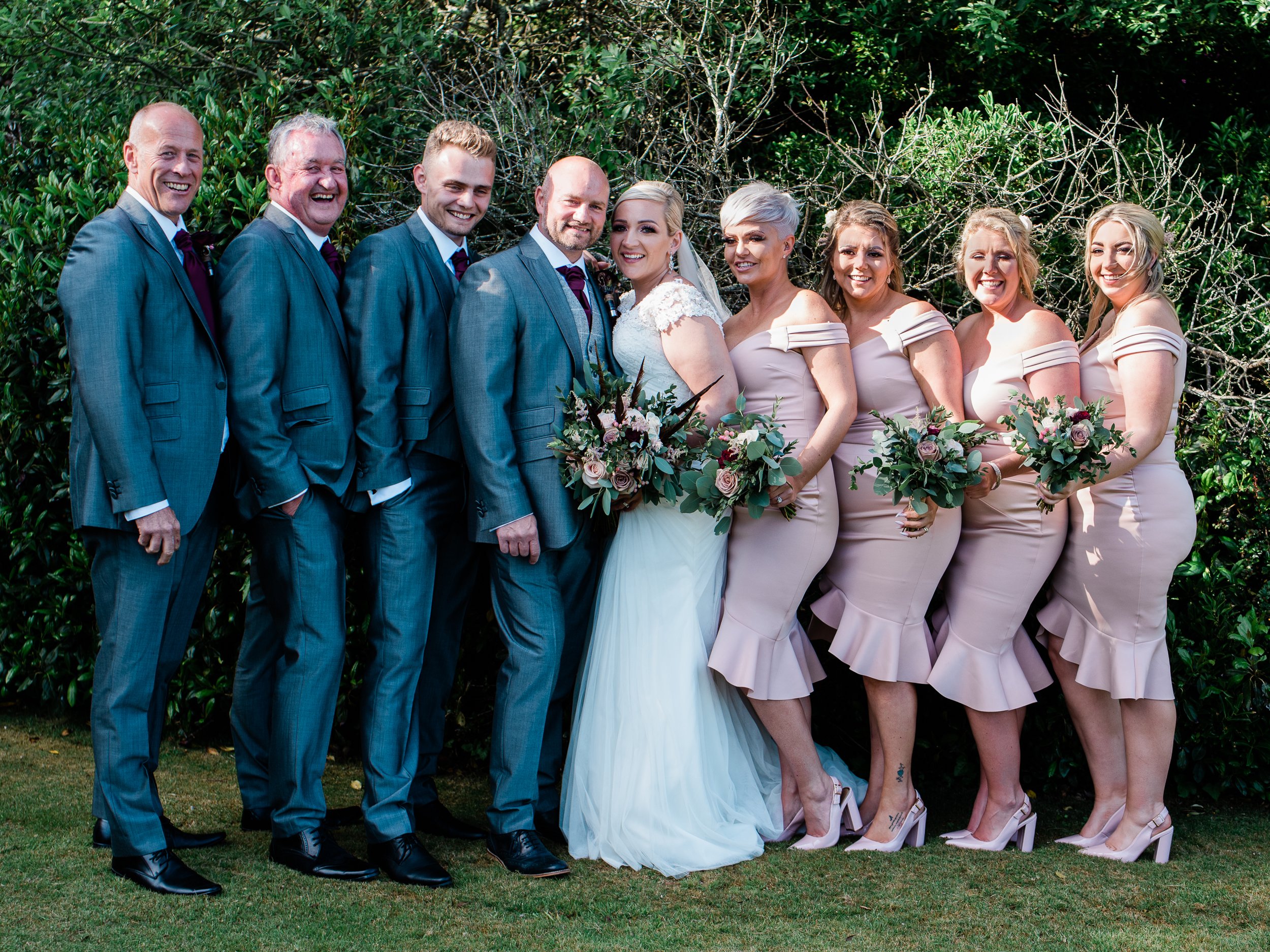 A wedding party outdoors with nine people, including the bride and groom in the center, surrounded by bridesmaids and groomsmen, all dressed formally, standing in front of greenery.