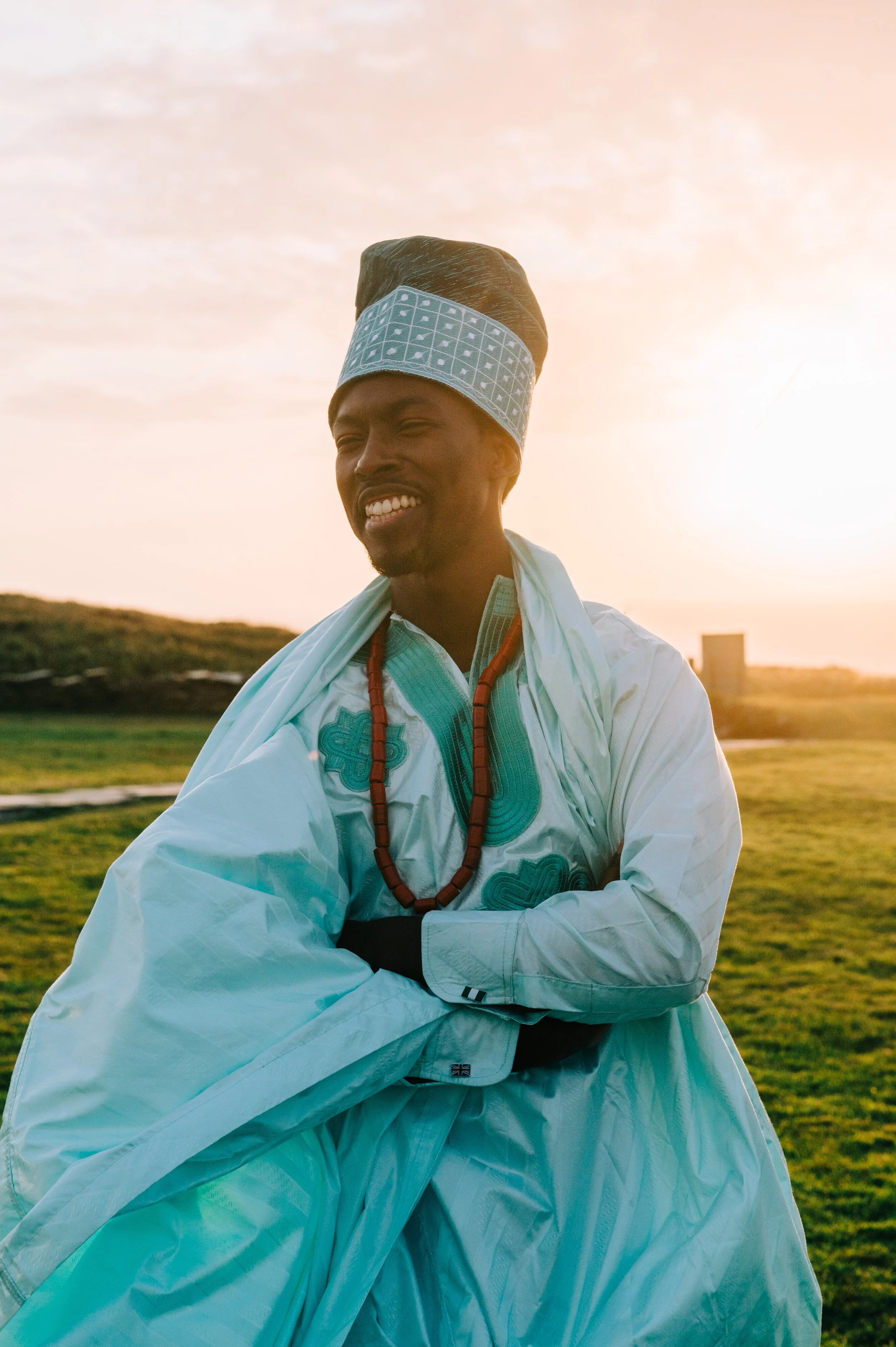A man wearing traditional Ethiopian attire, including a white robe and a tall hat with blue embroidery, standing outdoors at sunset with a grassy landscape in the background.