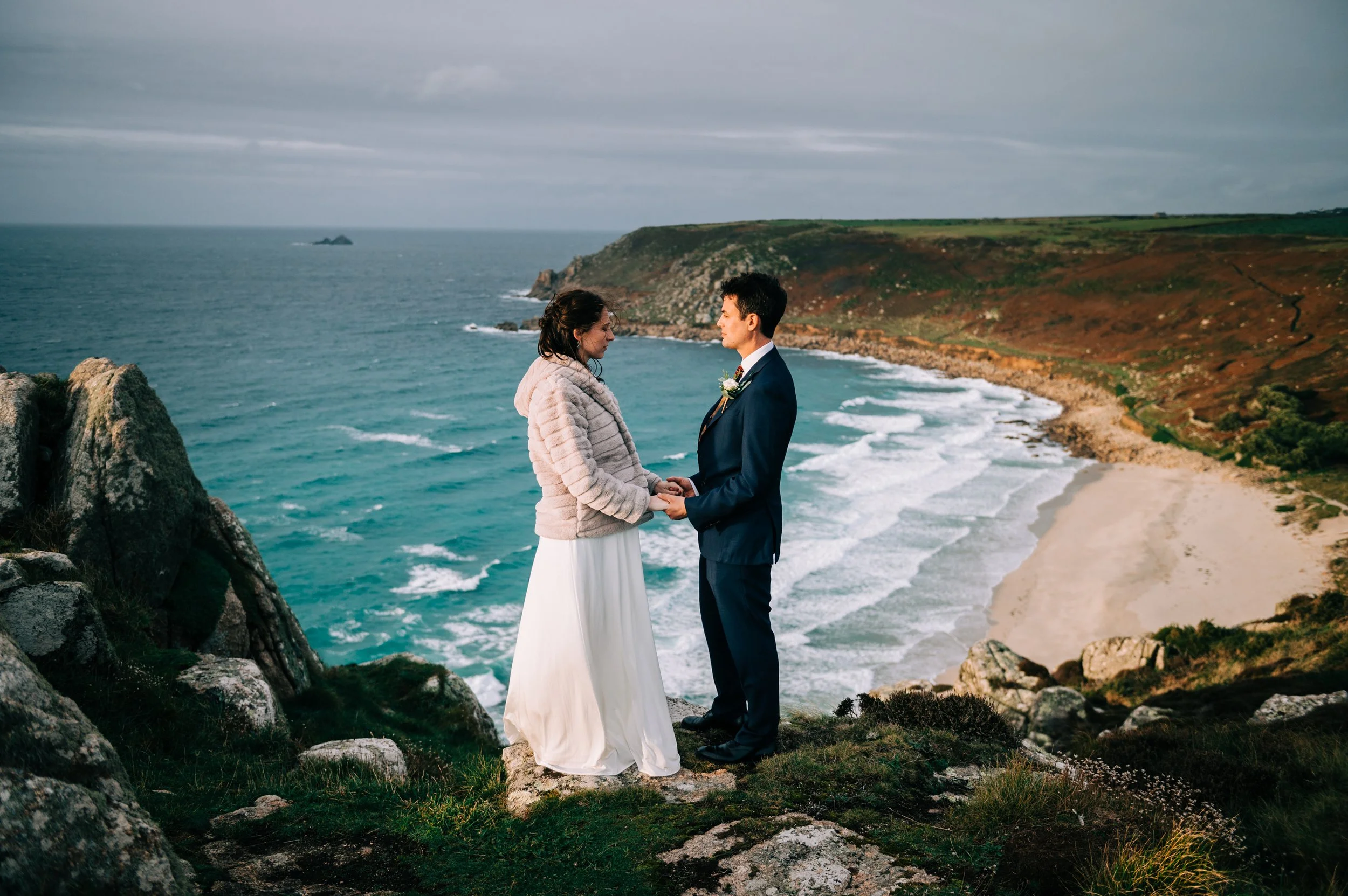 A couple in wedding attire holding hands and facing each other on a cliff with a view of a beach, ocean, and green hills in the background.
