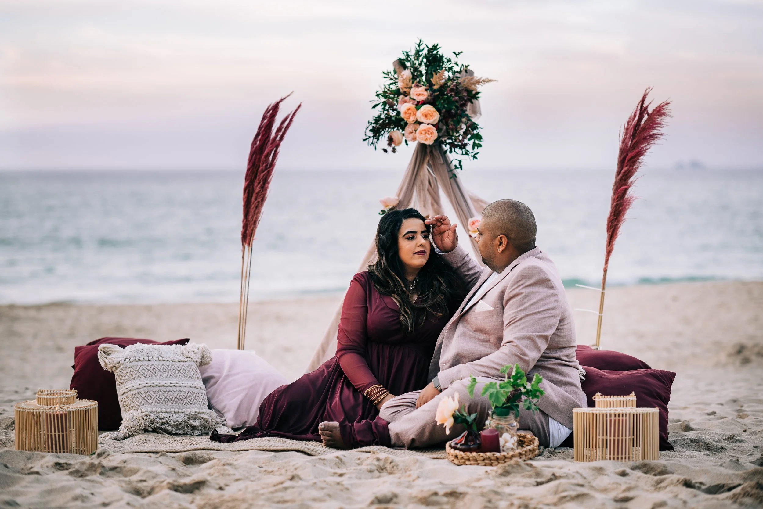 Couple sitting on a beach during sunset, with decorative pillows, flowers, and a floral arch behind them for a romantic scene.