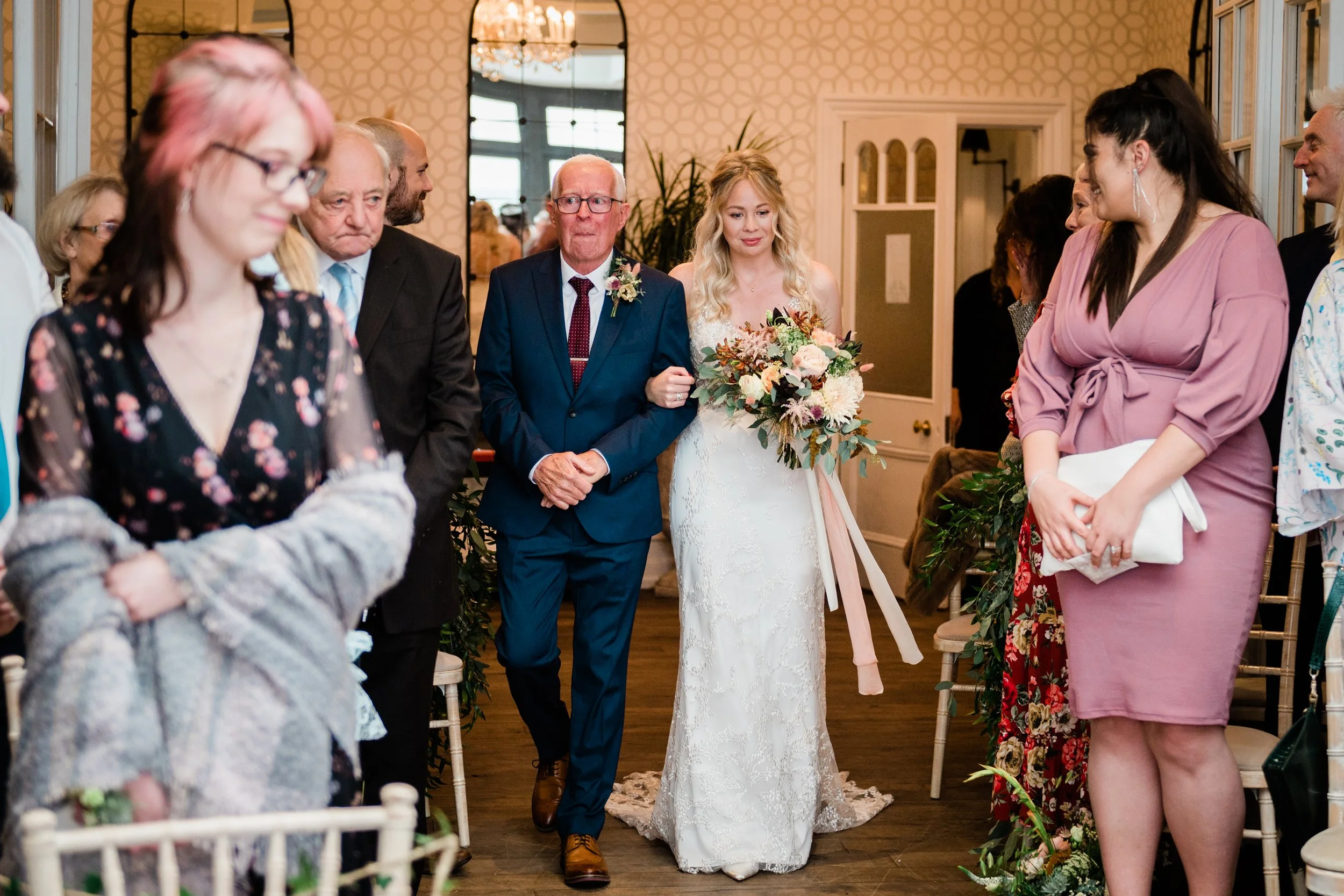 A bride walking down the aisle with her father during a wedding ceremony in a decorated indoor venue.