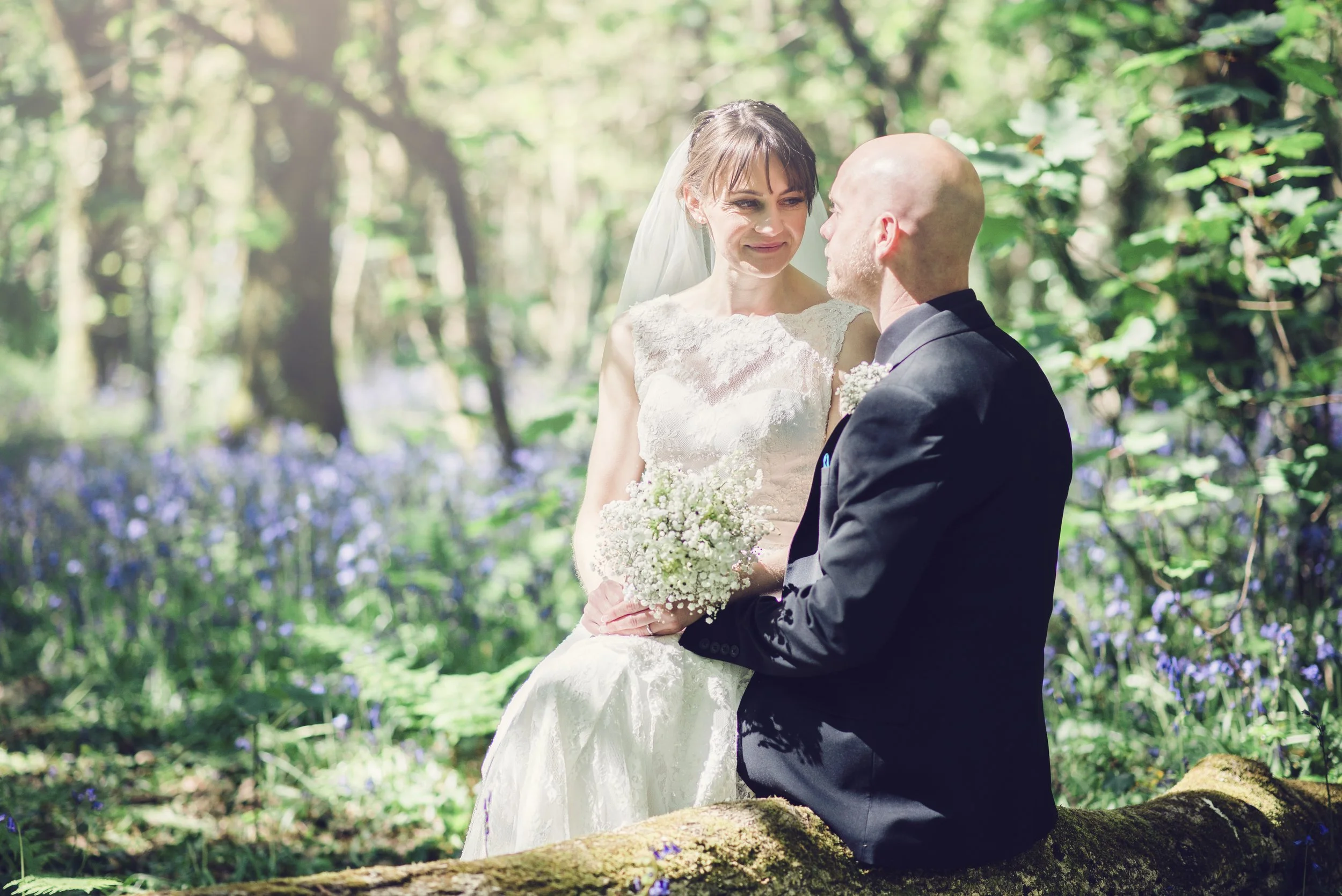 A bride and groom in a forest with sunlight shining through trees, the bride holding a bouquet and wearing a lace wedding dress, the groom dressed in a suit, sitting on a log with greenery and purple flowers surrounding them.