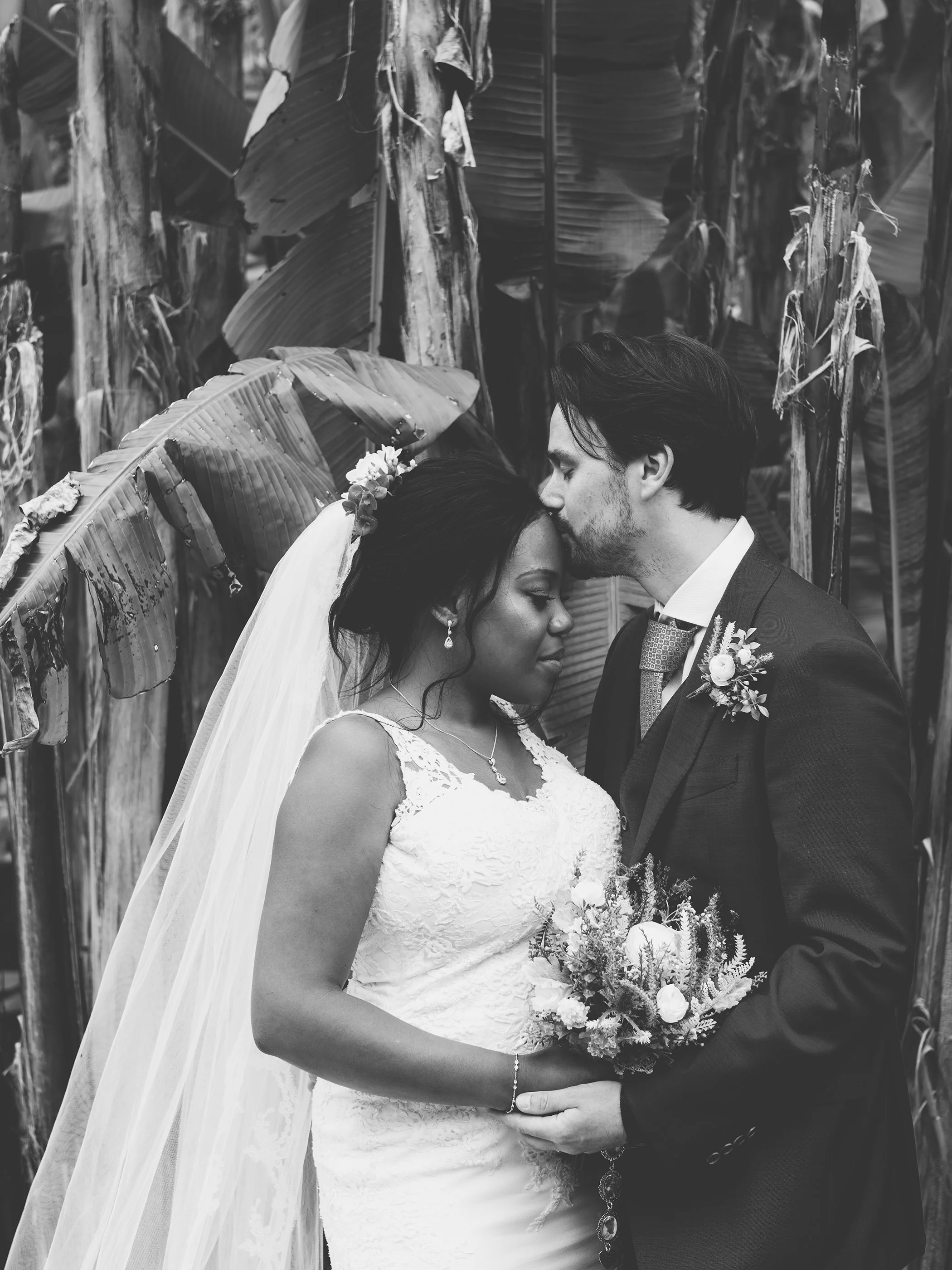 Black and white photo of a bride and groom embracing, with the groom kissing the bride's forehead. The bride is holding a bouquet and wearing a wedding dress with a veil, while the groom is in a suit with a boutonniere. They are surrounded by large l
