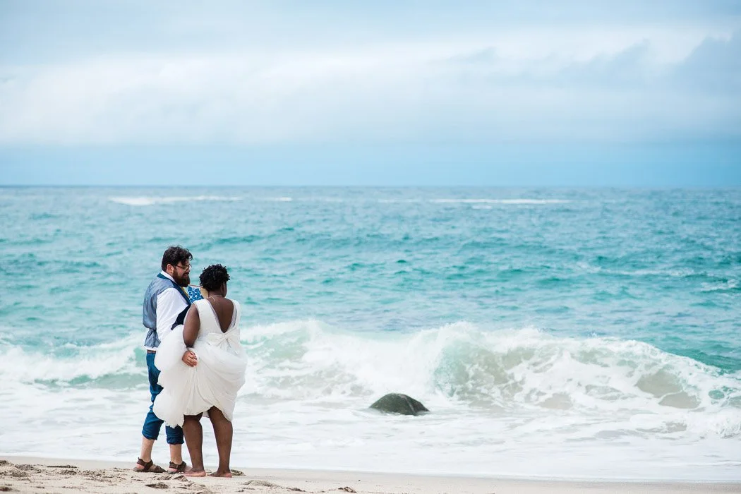 A couple dressed in wedding attire standing on a beach near the water, with waves and a cloudy sky in the background.
