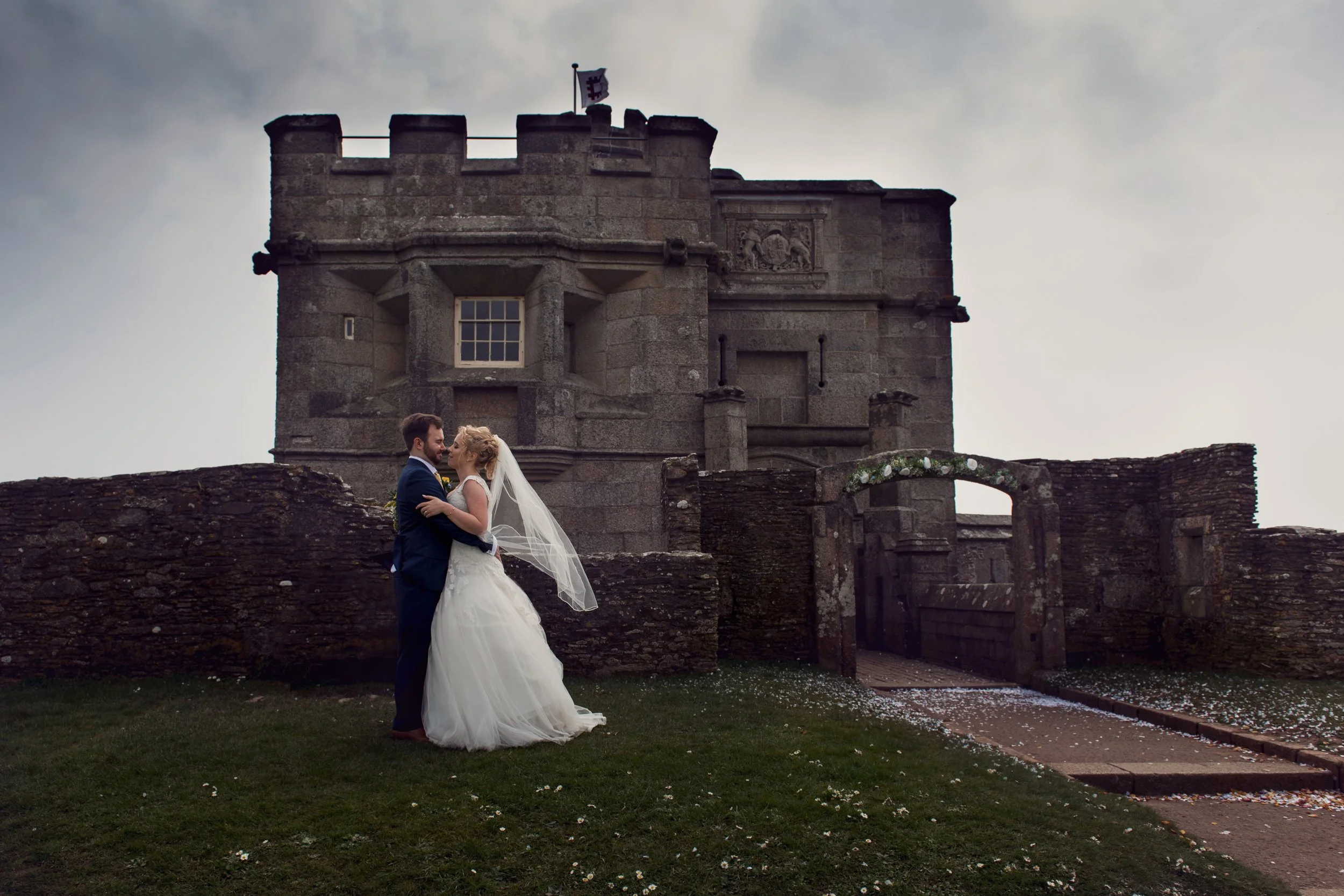 A bride and groom stand close together in front of a castle, sharing a kiss. The bride is wearing a white wedding dress and veil, and the groom is in a navy suit. The sky is overcast.