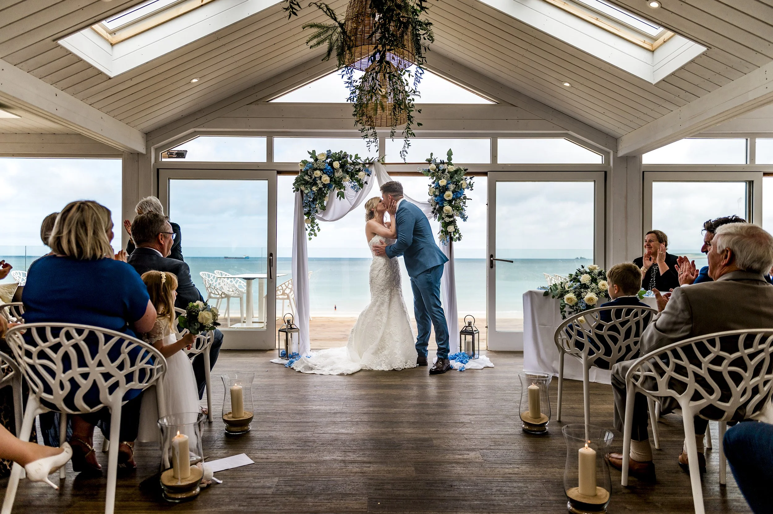 A wedding ceremony with a couple kissing under an arch decorated with flowers, overlooking the beach with the ocean in the background. Guests are seated on either side, clapping and celebrating.