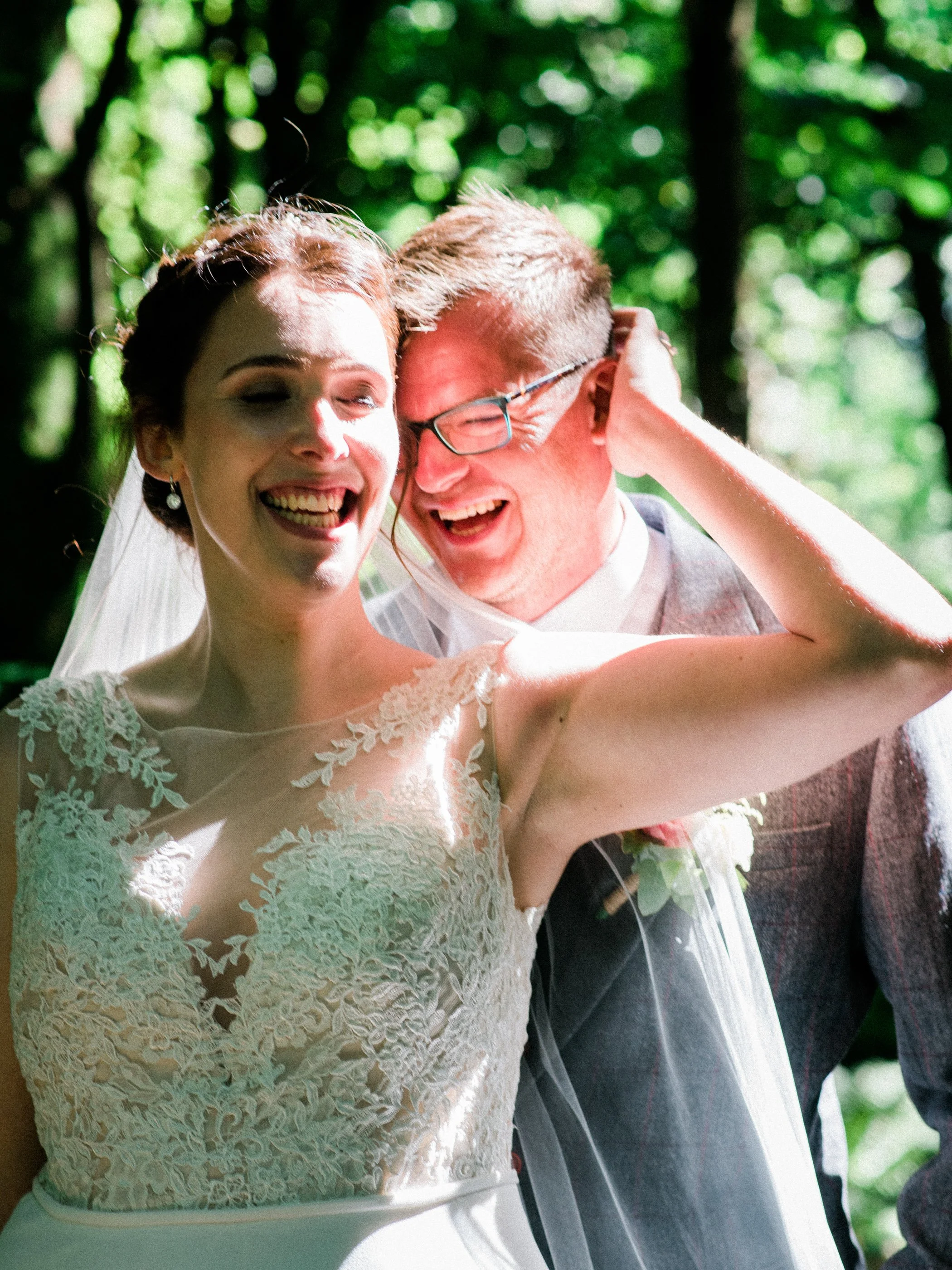 A bride and groom smiling and laughing together outdoors during their wedding, with trees and sunlight in the background.