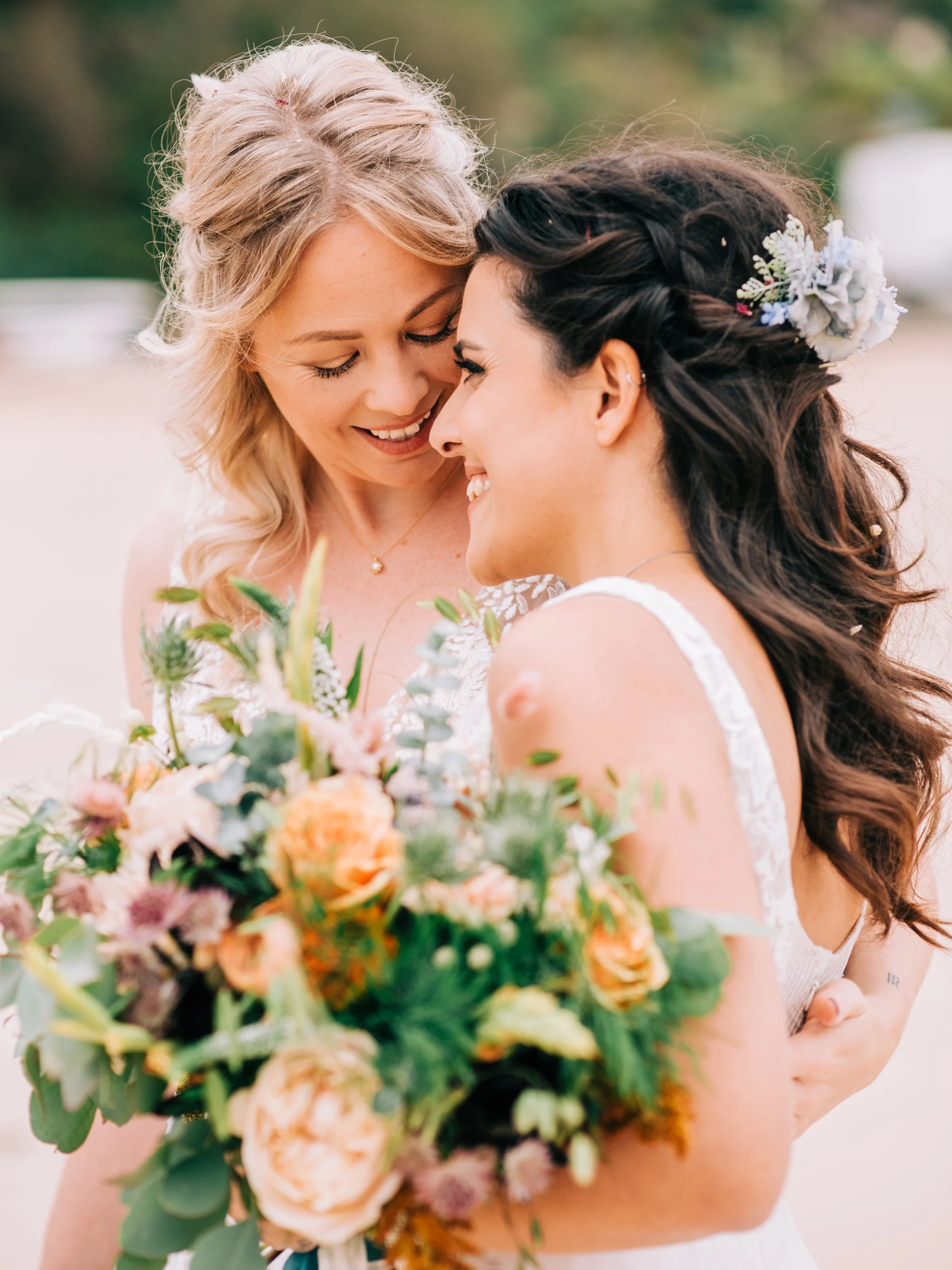 Two women, one with blonde hair and one with dark hair, sharing an intimate moment, holding a bouquet of colorful flowers, outdoors on a bright day.