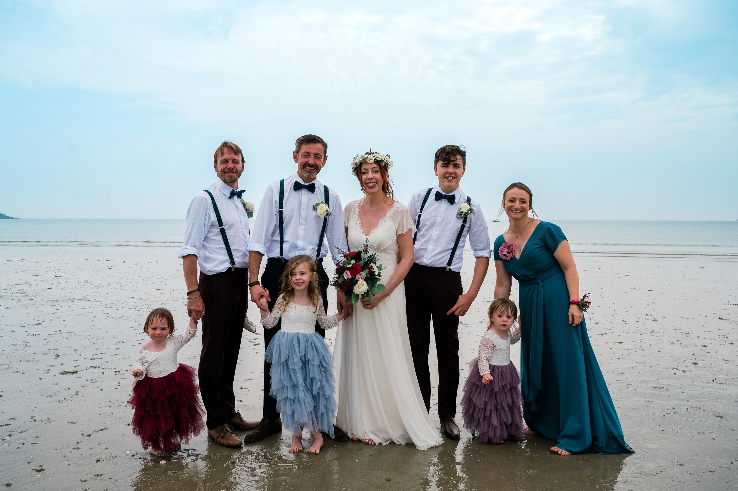A group of people at a beach for a wedding, including a bride in a white dress holding a bouquet, five adults, and three young children in colorful dresses.