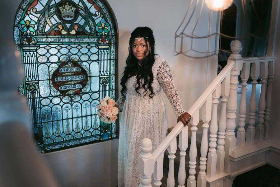 A woman in a white lace dress holding a bouquet of pink and white roses standing on a staircase next to a stained glass window with a crown emblem.