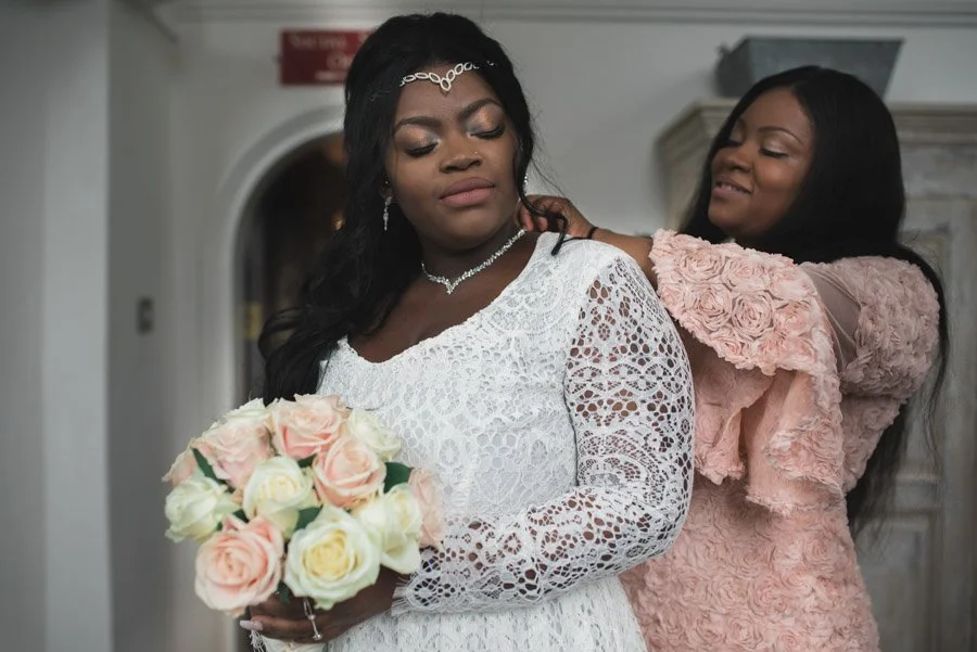 Two women, one dressed as a bride holding a bouquet of pink and white roses, the other woman is adjusting the bride's dress and wearing a pink lace dress, standing in an indoor setting.