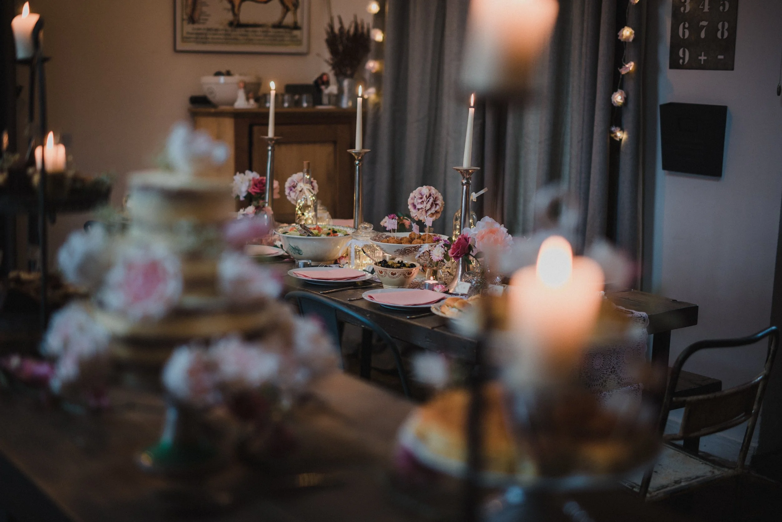 A decorated dining table set for a meal with candles, flowers, bowls of food, and plates. The background includes a wooden console, a painting, and curtains.