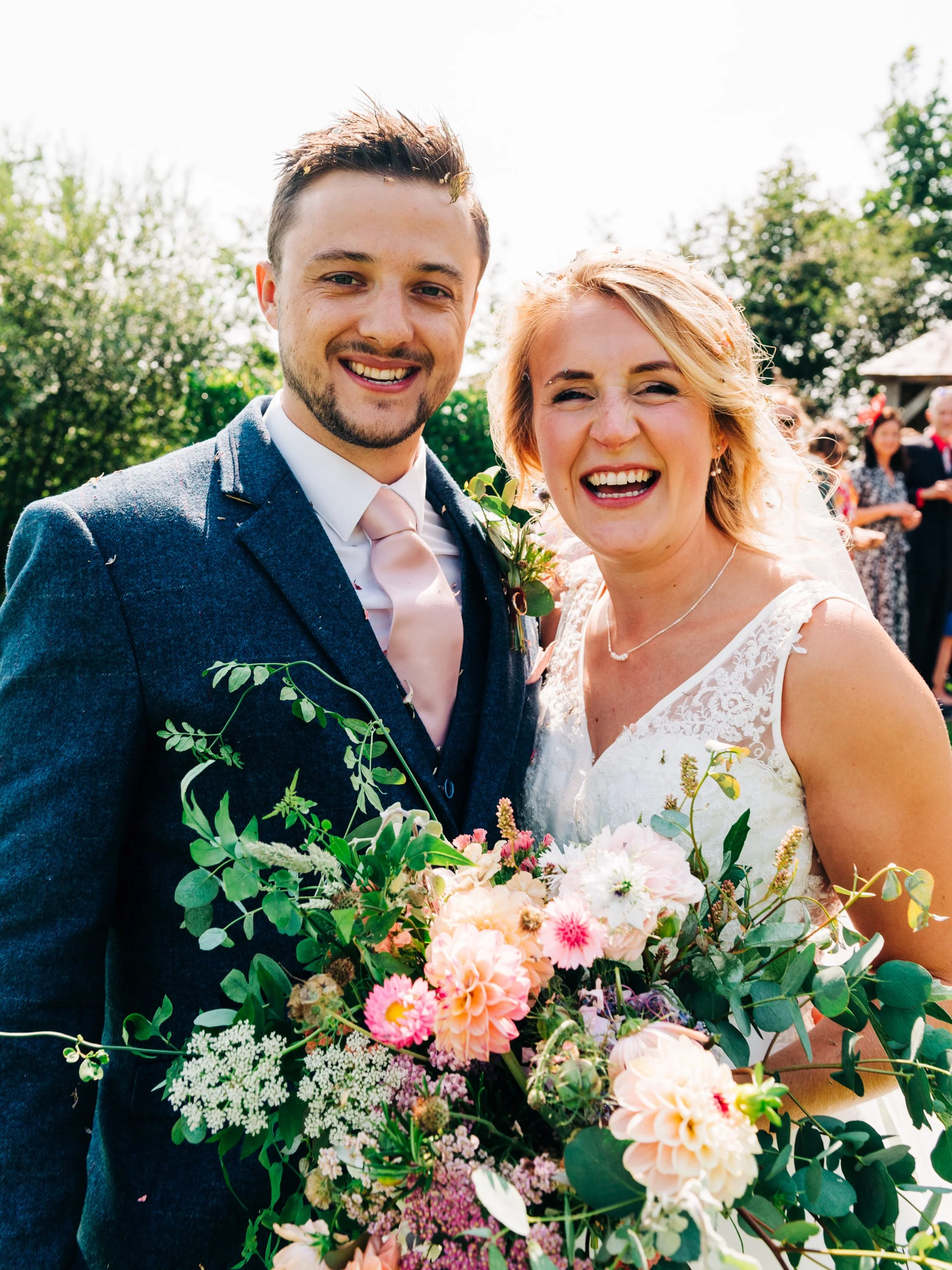 A smiling bride and groom in wedding attire outdoors, holding a large bouquet of flowers, with guests in the background.