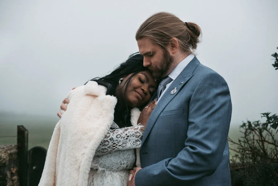 A woman and a man sharing a tender moment outdoors, with the woman resting her head on the man's chest and the man holding her gently. The woman has long dark hair and is wearing a white dress and a fur shawl, while the man has long blond hair tied b