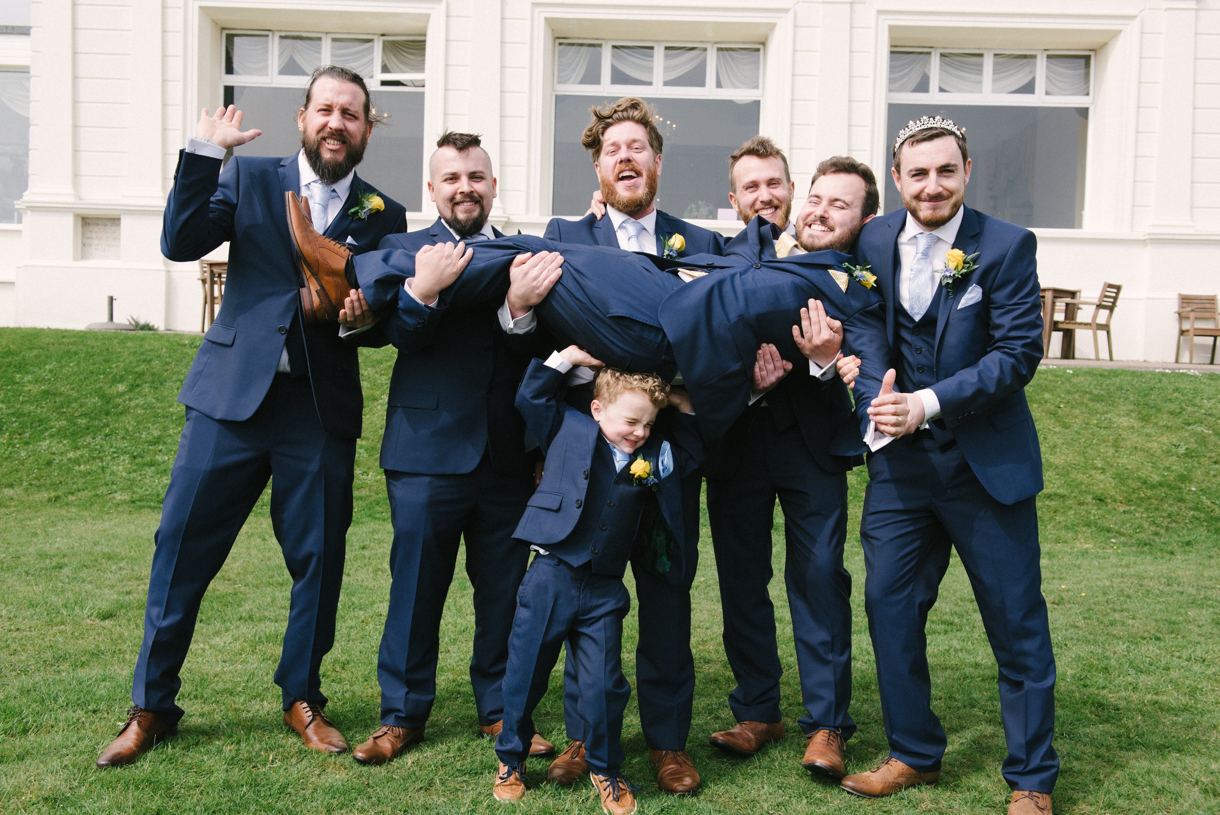A group of men in suits, with one man dressed as royalty, lifting another man horizontally in a celebratory pose on a grassy lawn in front of a white building with large windows.