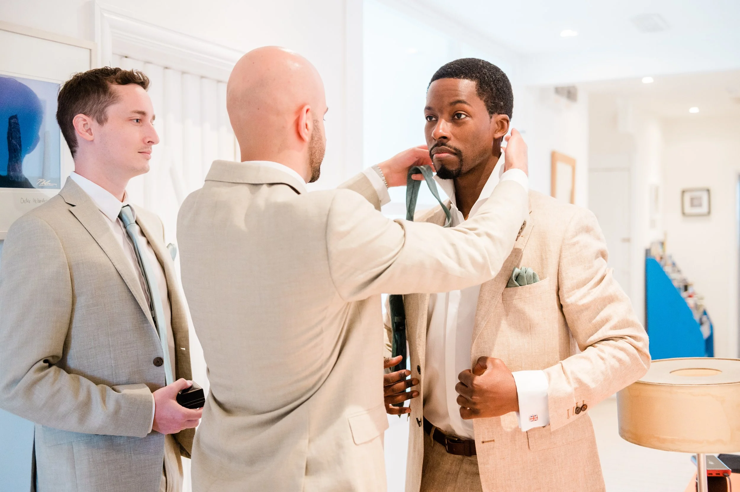 Three men in beige suits standing indoors, one is attaching a green tie around the neck of another man, who is holding his jacket lapel with one hand, in a well-lit room with artwork and books in the background.