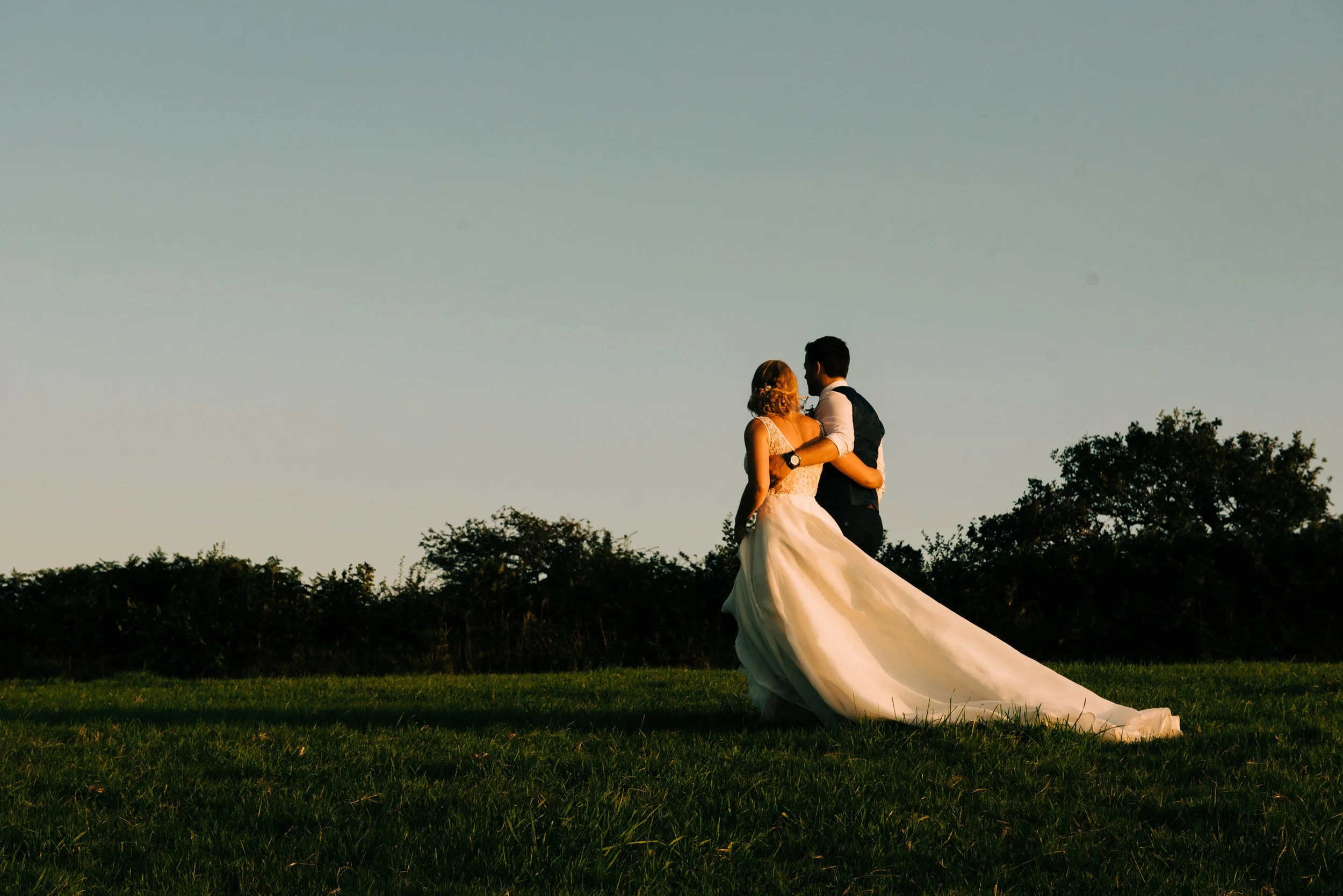 A bride and groom dancing outdoors on a grassy field during sunset, with trees in the background.