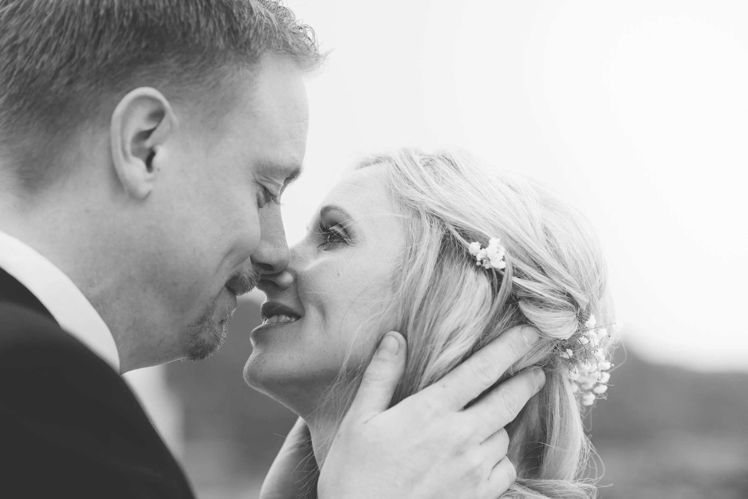 A black and white photo of a couple in wedding attire, close-up, with faces near each other, smiling softly, outdoors with a blurred background.
