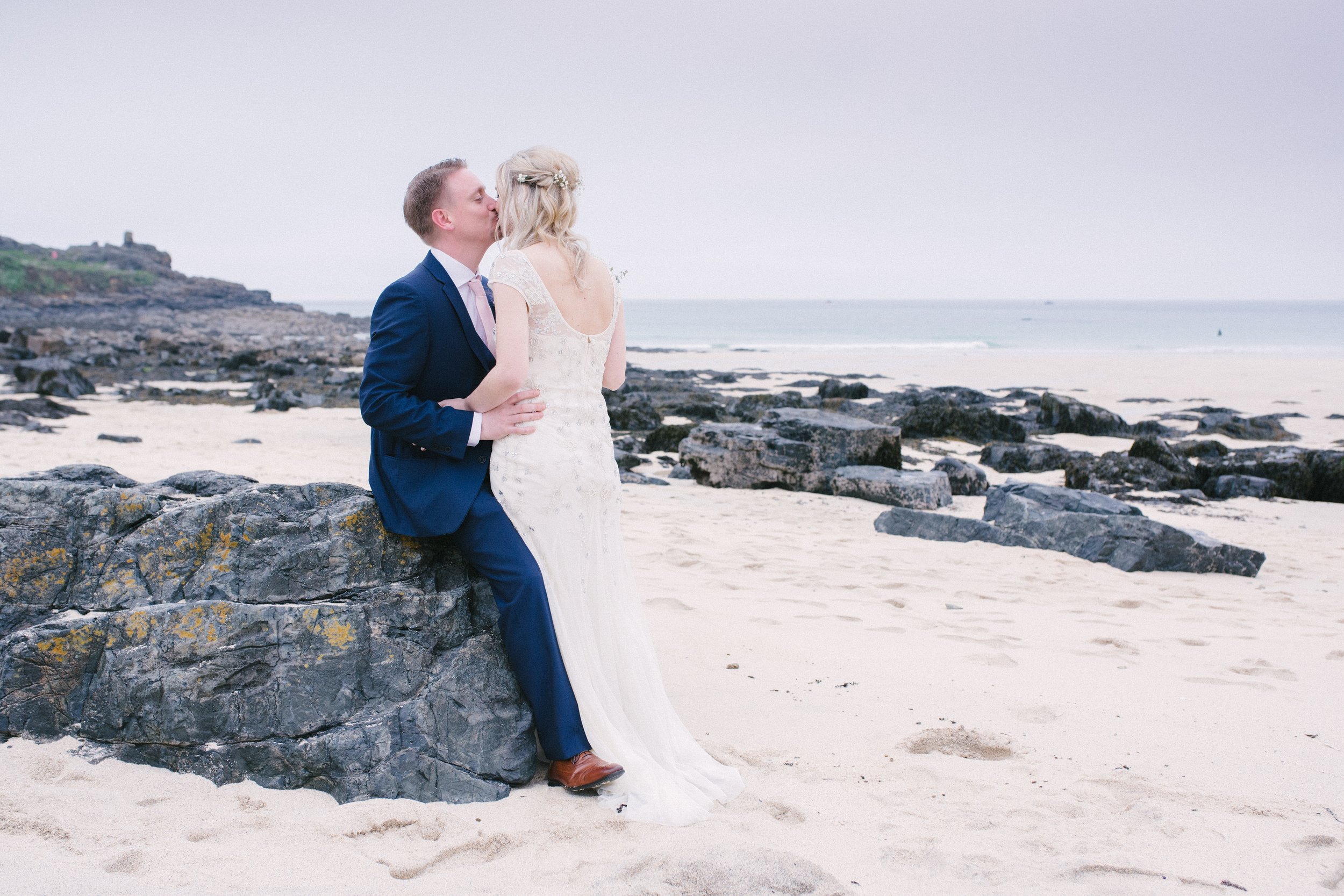 A newlywed couple in wedding attire sharing a kiss on a rocky beach.