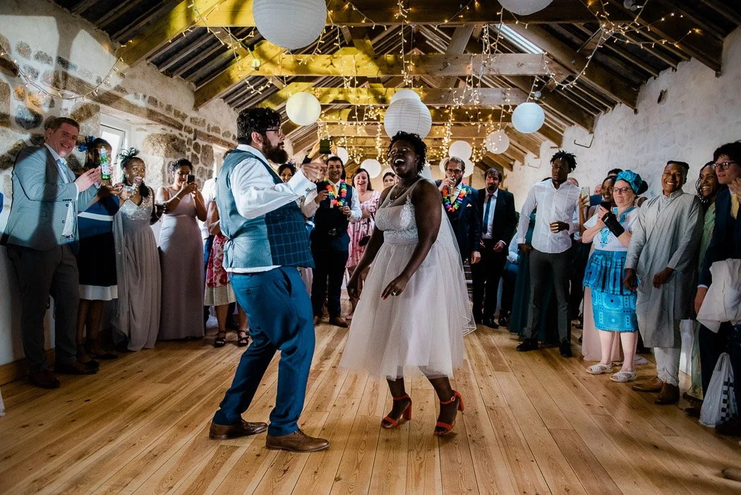 Couple dancing at a wedding reception with guests watching in a rustic hall decorated with paper lanterns and string lights.
