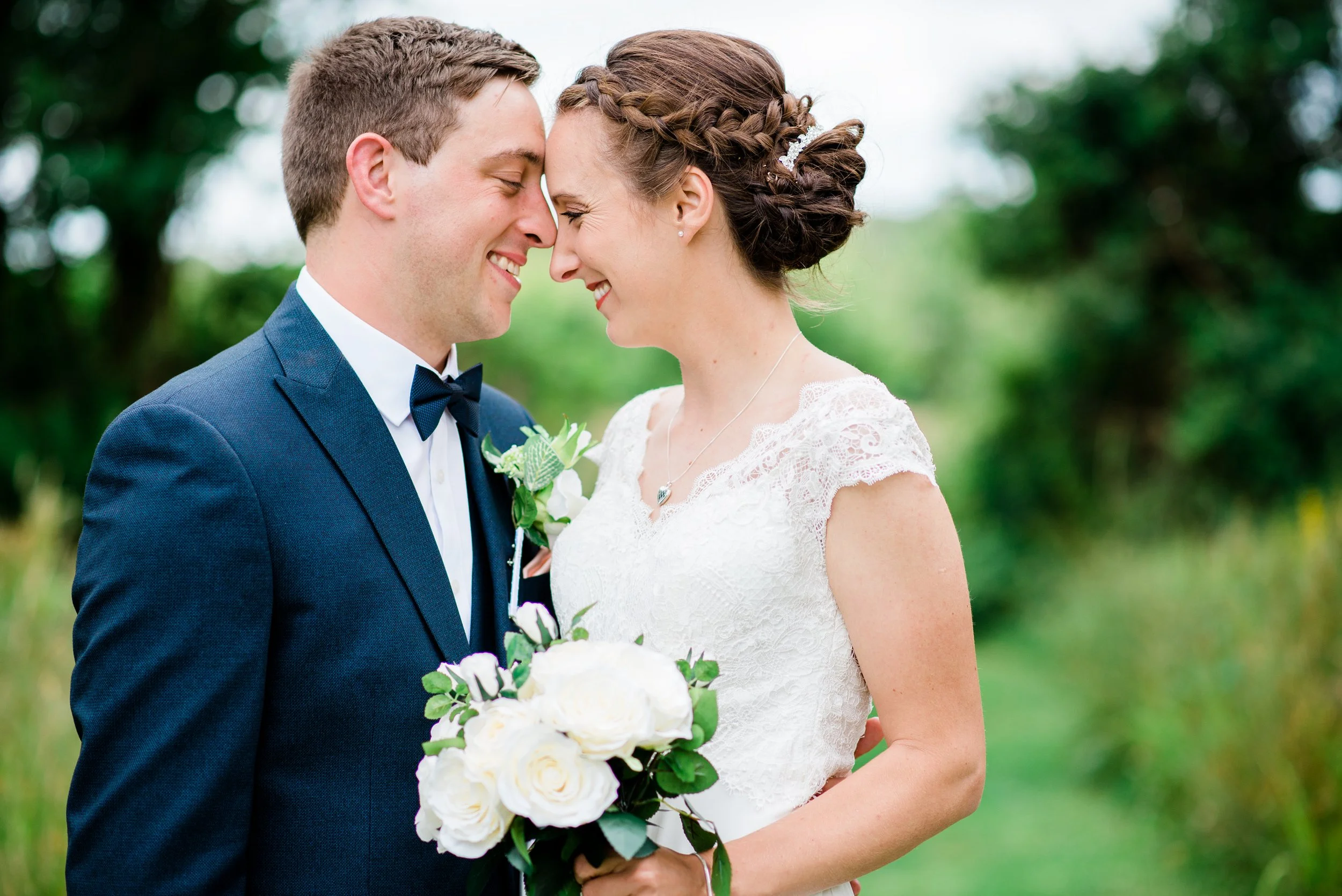 Bride and groom with foreheads touching, smiling, outdoors with greenery in the background, bride holding a bouquet of white roses.