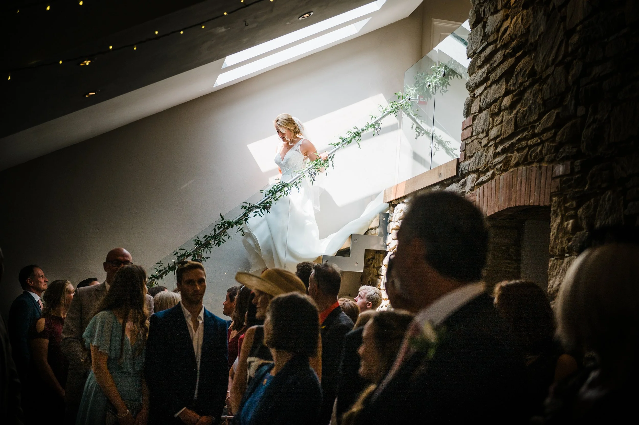 A bride in a white wedding dress walking down a staircase decorated with greenery, with guests watching below in a dimly lit indoor setting.