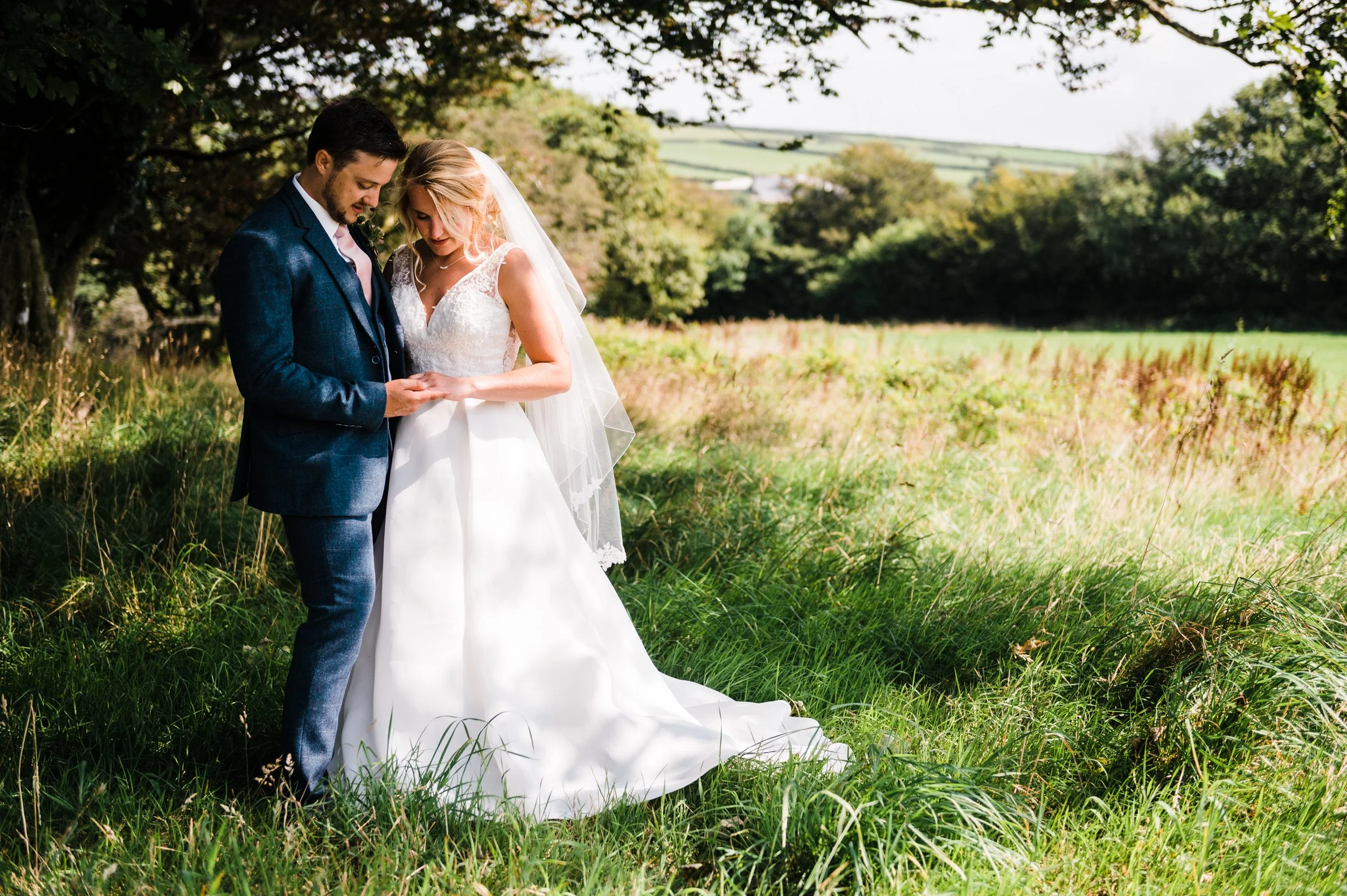 A bride and groom standing together in a grassy field holding hands, with trees and distant hills in the background, on a sunny day.
