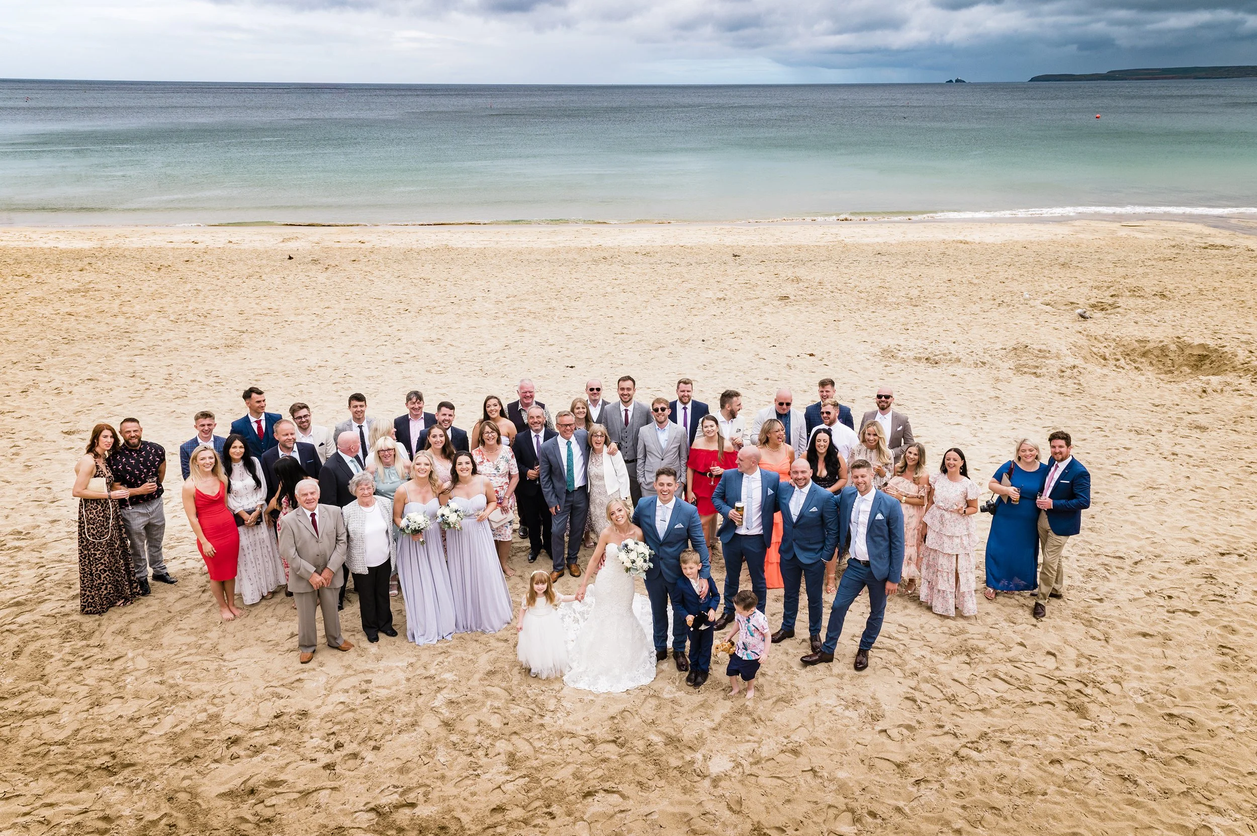 A large group of people celebrating a wedding on a sandy beach near the ocean. The bride and groom are in the center, with the bride wearing a white wedding dress and holding a bouquet, and the groom in a blue suit. Other guests are dressed in formal