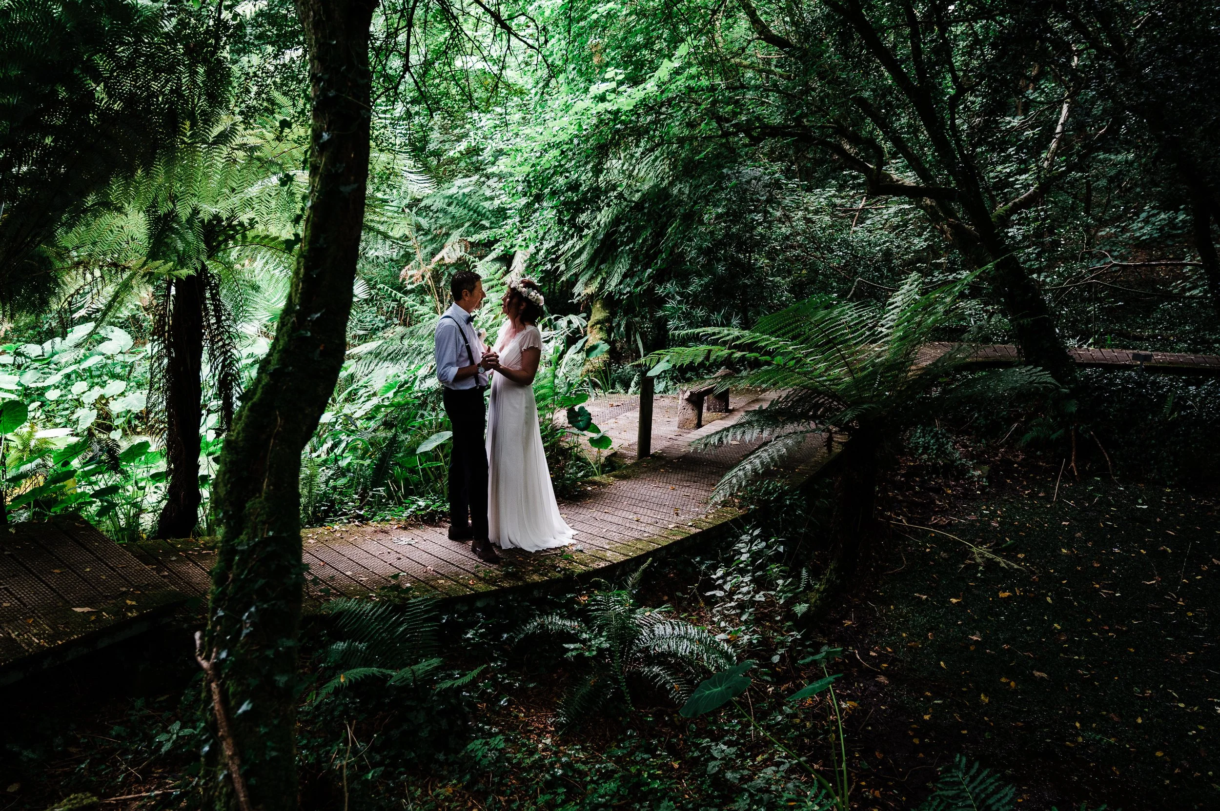 A couple dressed in wedding attire stands on a wooden pathway in a lush green forest, holding hands and looking into each other's eyes.
