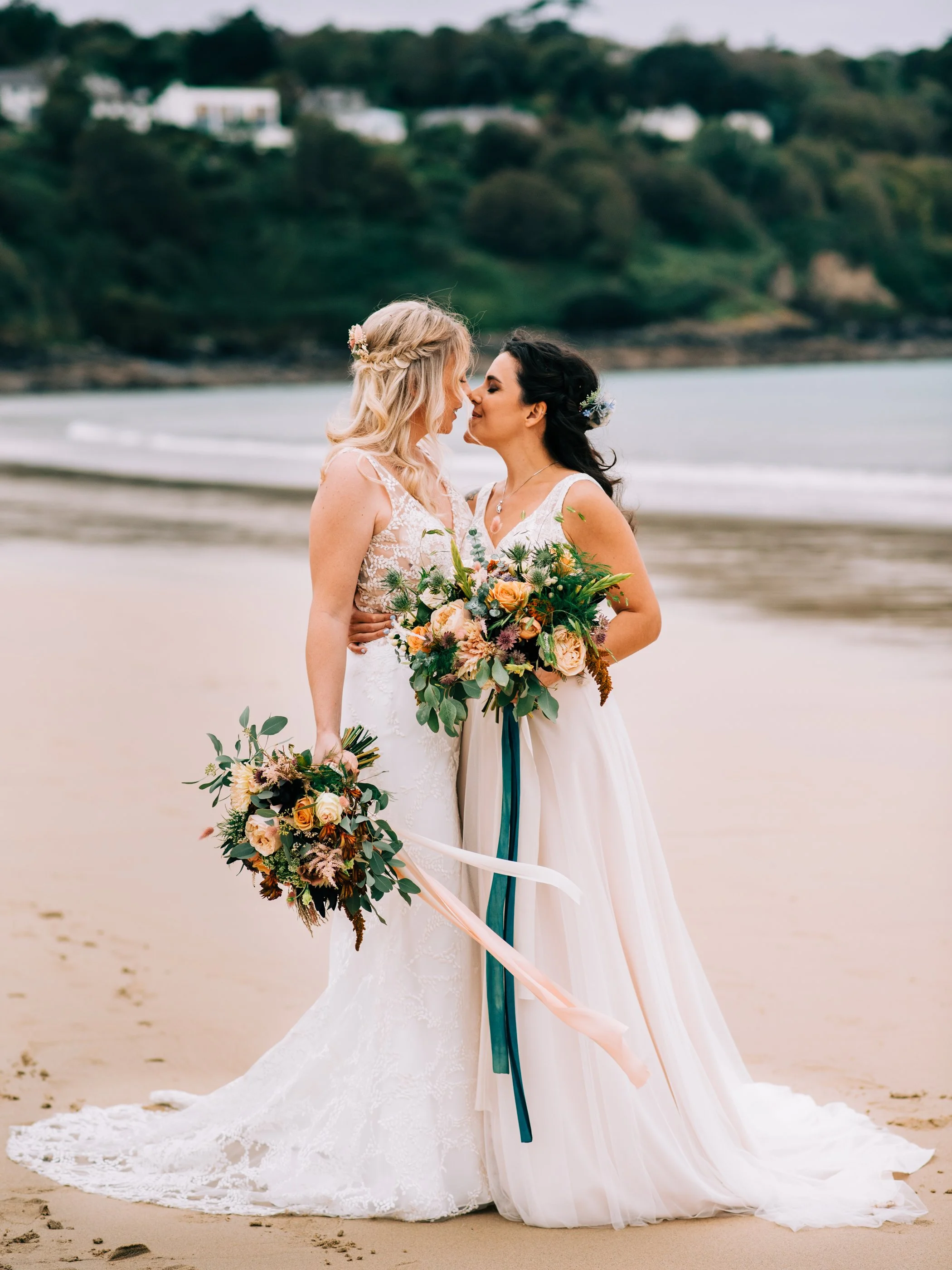 Two women in wedding dresses standing on a beach, holding bouquets, and touching foreheads in an embrace.