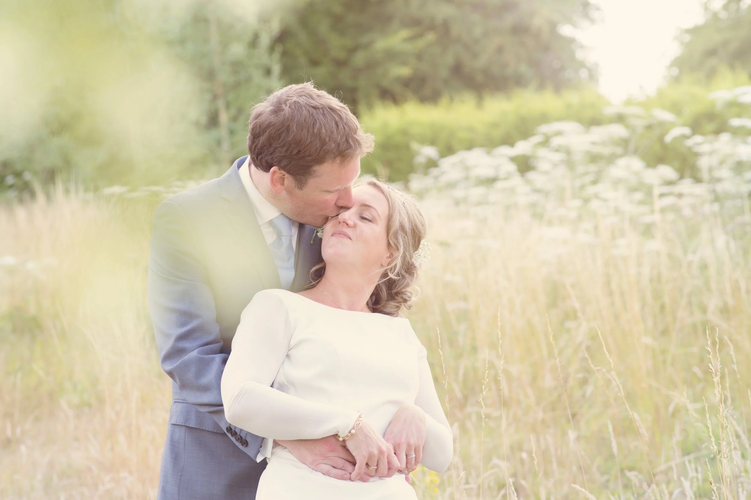 A man in a gray suit kisses a woman in a white dress on the forehead in a field of tall grass with trees in the background.