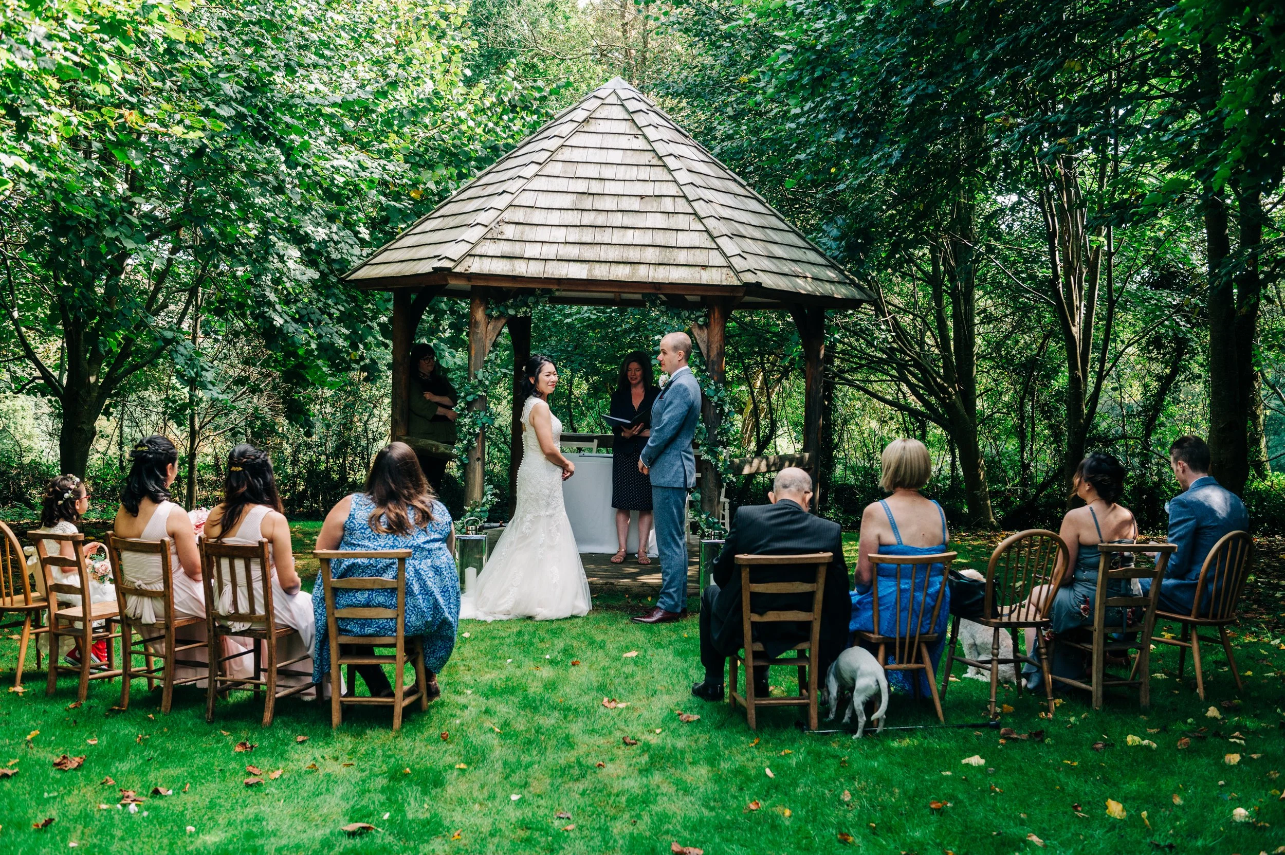 Outdoor wedding ceremony in a lush green garden with a small wooden pavilion, bride and groom exchanging vows, guests seated on chairs, some holding bouquets, a dog among the guests, and an officiant officiating the ceremony.