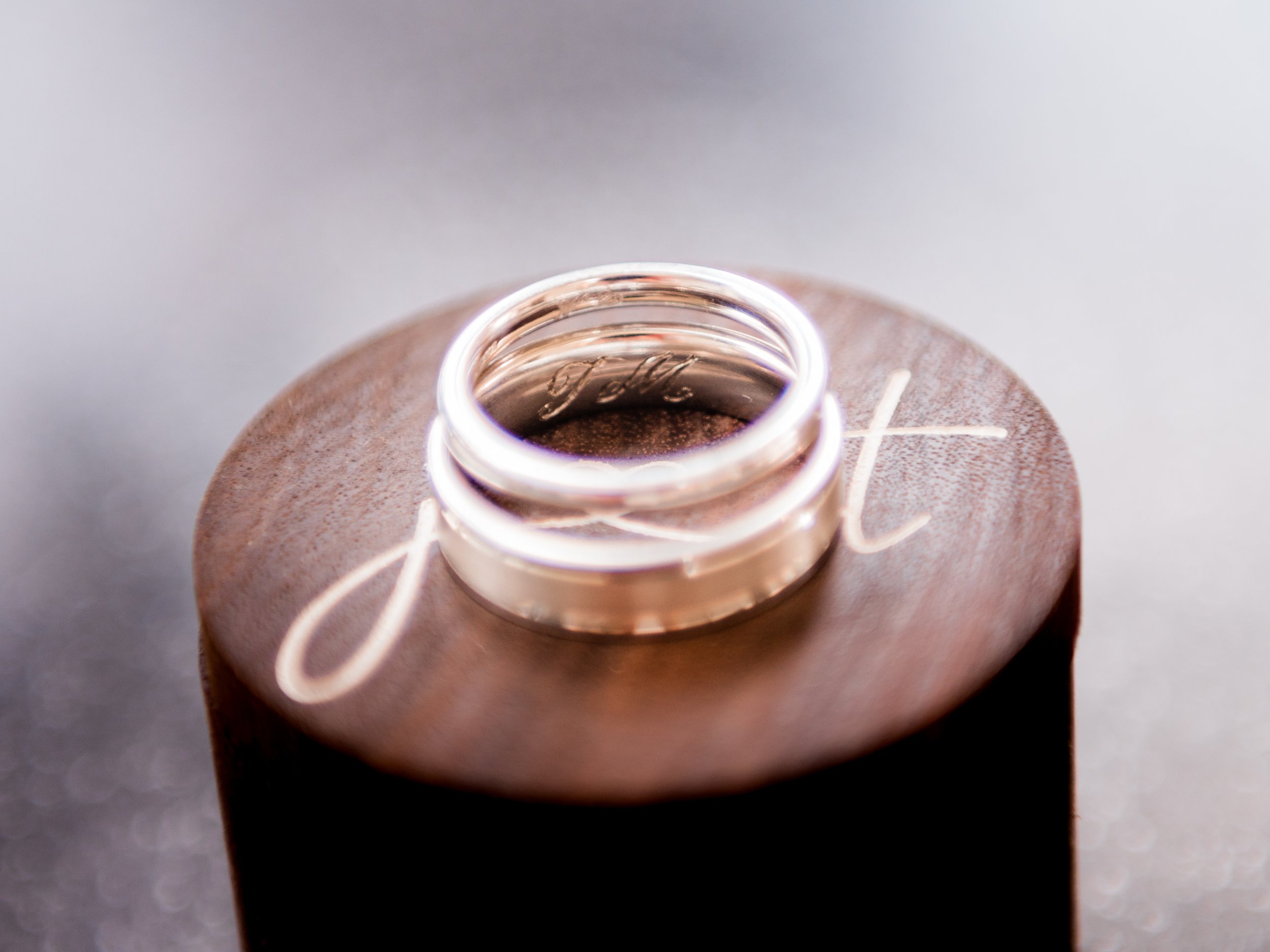 Close-up of two silver rings on a wooden surface with white engraved text, illuminated with soft lighting.