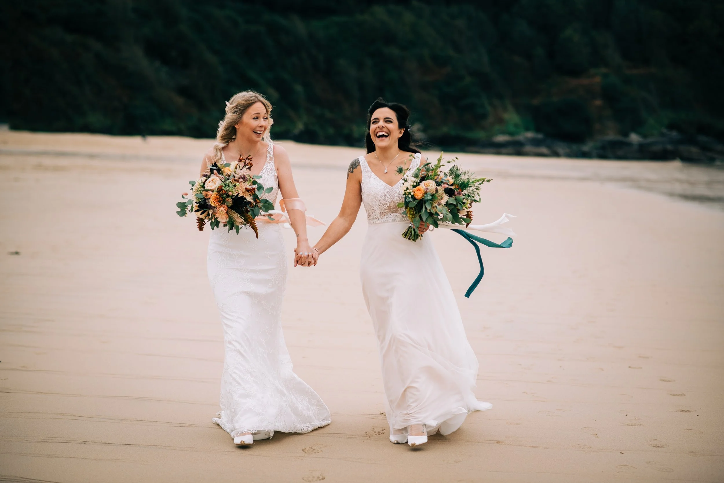 Two women in wedding dresses holding hands and bouquets, walking on a sandy beach with greenery in the background, smiling and enjoying the moment.