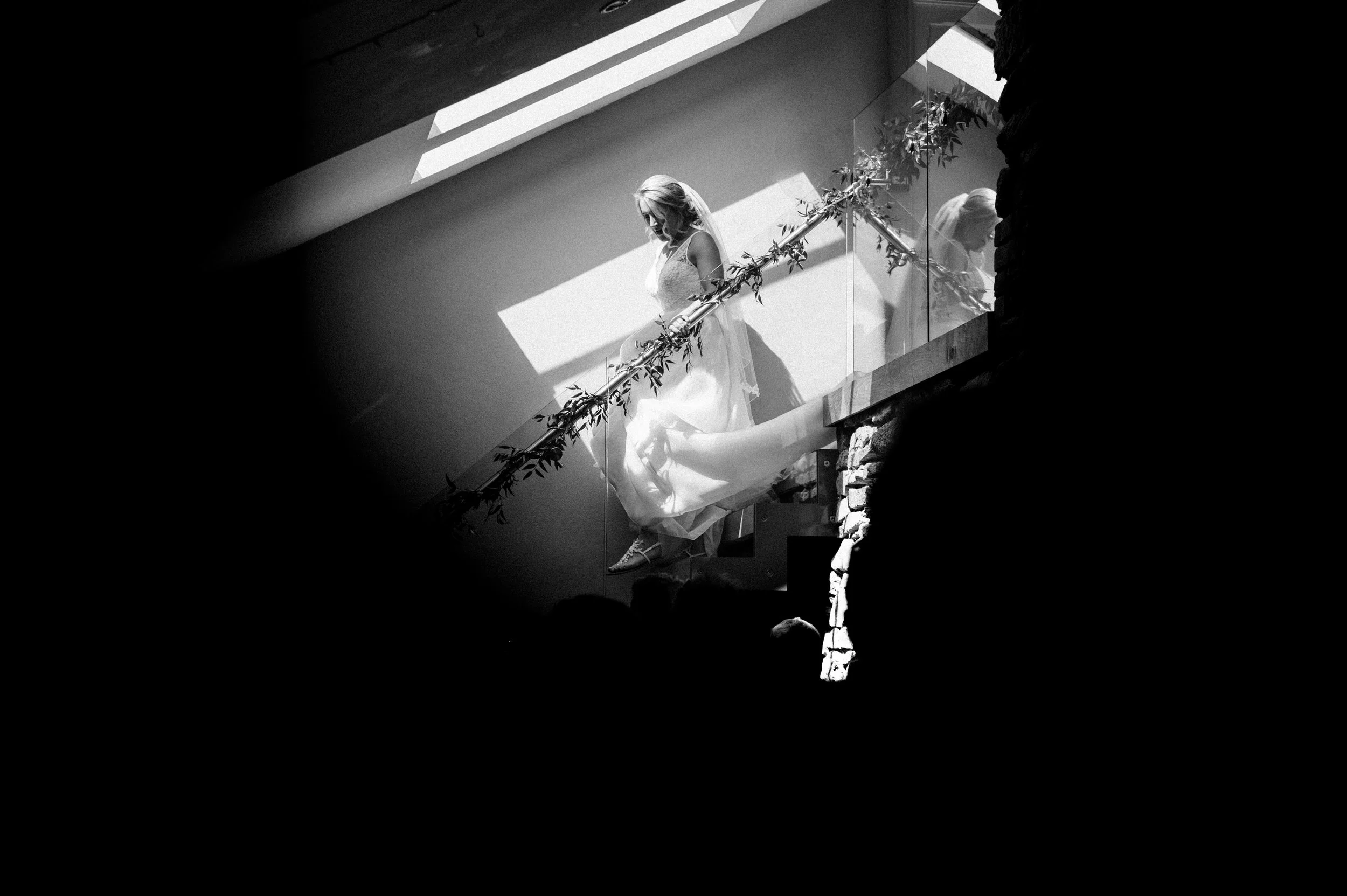 A bride in a wedding dress walking down stairs illuminated by natural light, with her reflection visible in the glass railing.