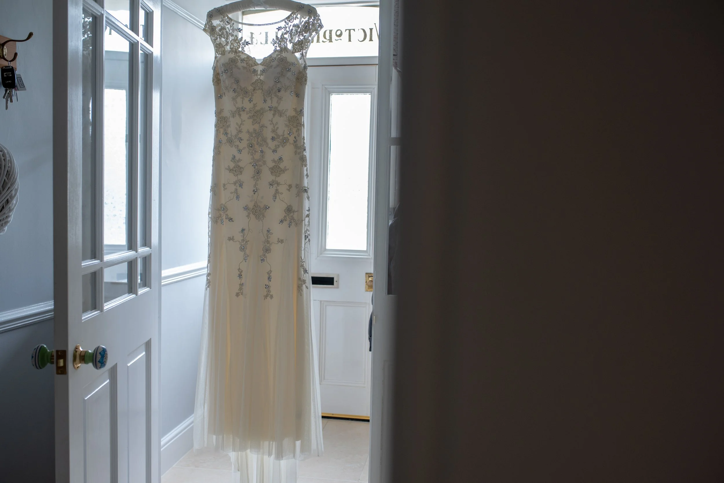 A white wedding dress with lace and bead embellishments hanging on a door in a well-lit entrance area.