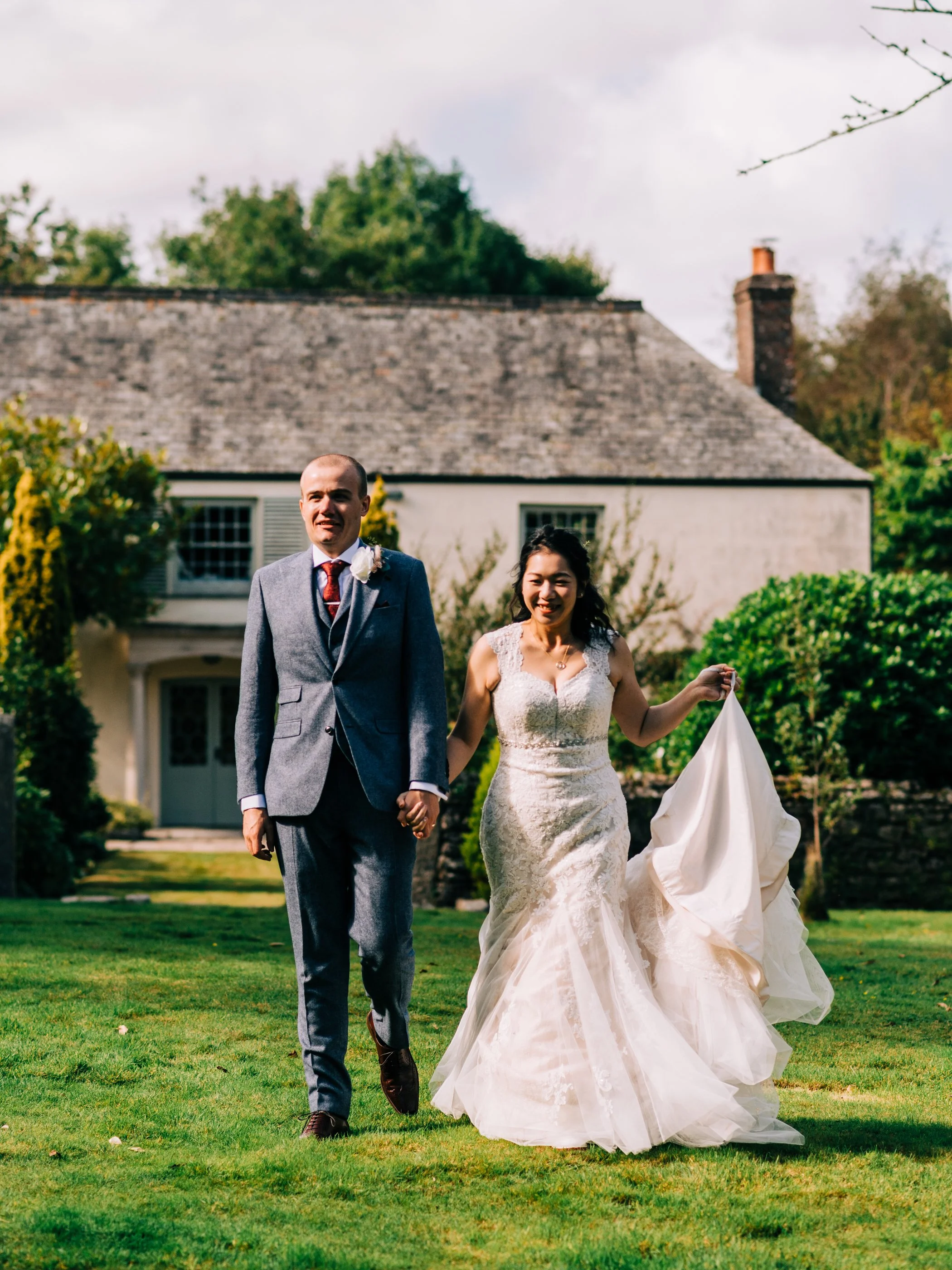 A newlywed couple walking hand in hand on a lawn in front of a house, smiling, with the bride holding up her wedding dress.