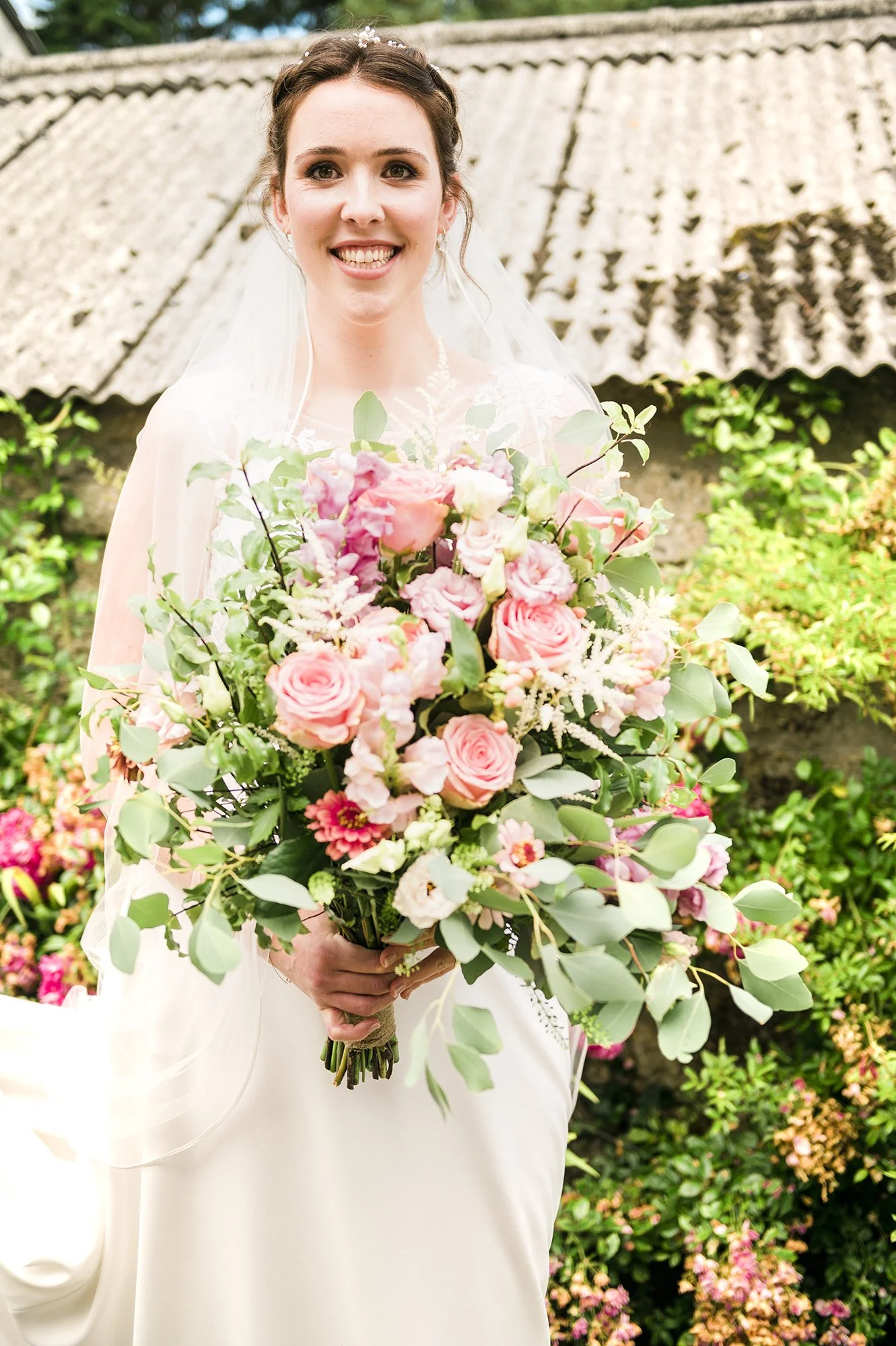 A bride in a white wedding dress holding a large bouquet of pink and white flowers, standing outdoors with greenery and an old roof behind her.