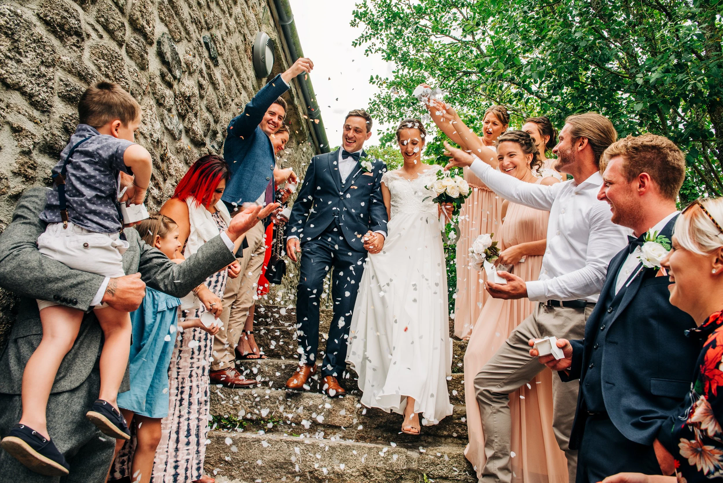 Bride and groom walking down stone steps surrounded by smiling guests throwing confetti during a wedding celebration outdoors with green trees.