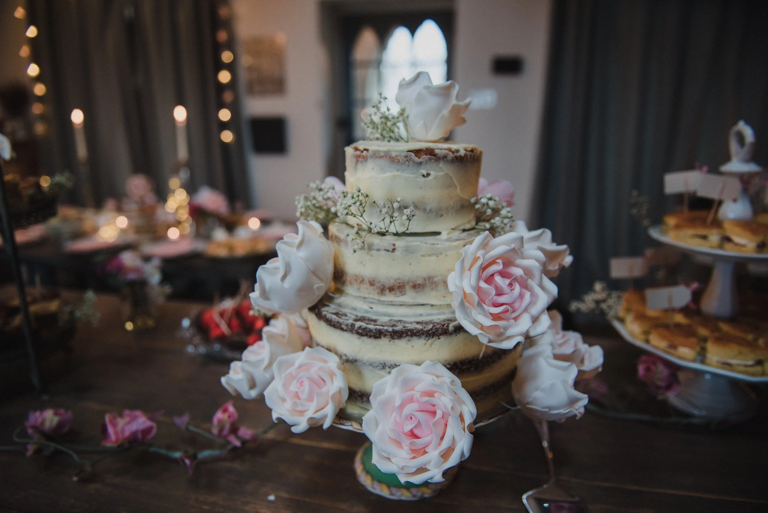 A rustic, semi-naked wedding cake decorated with white and pink artificial roses and small white flowers, placed on a glass cake stand. In the background, a decorated table with candles and food, inside a softly lit room with dark curtains and large 