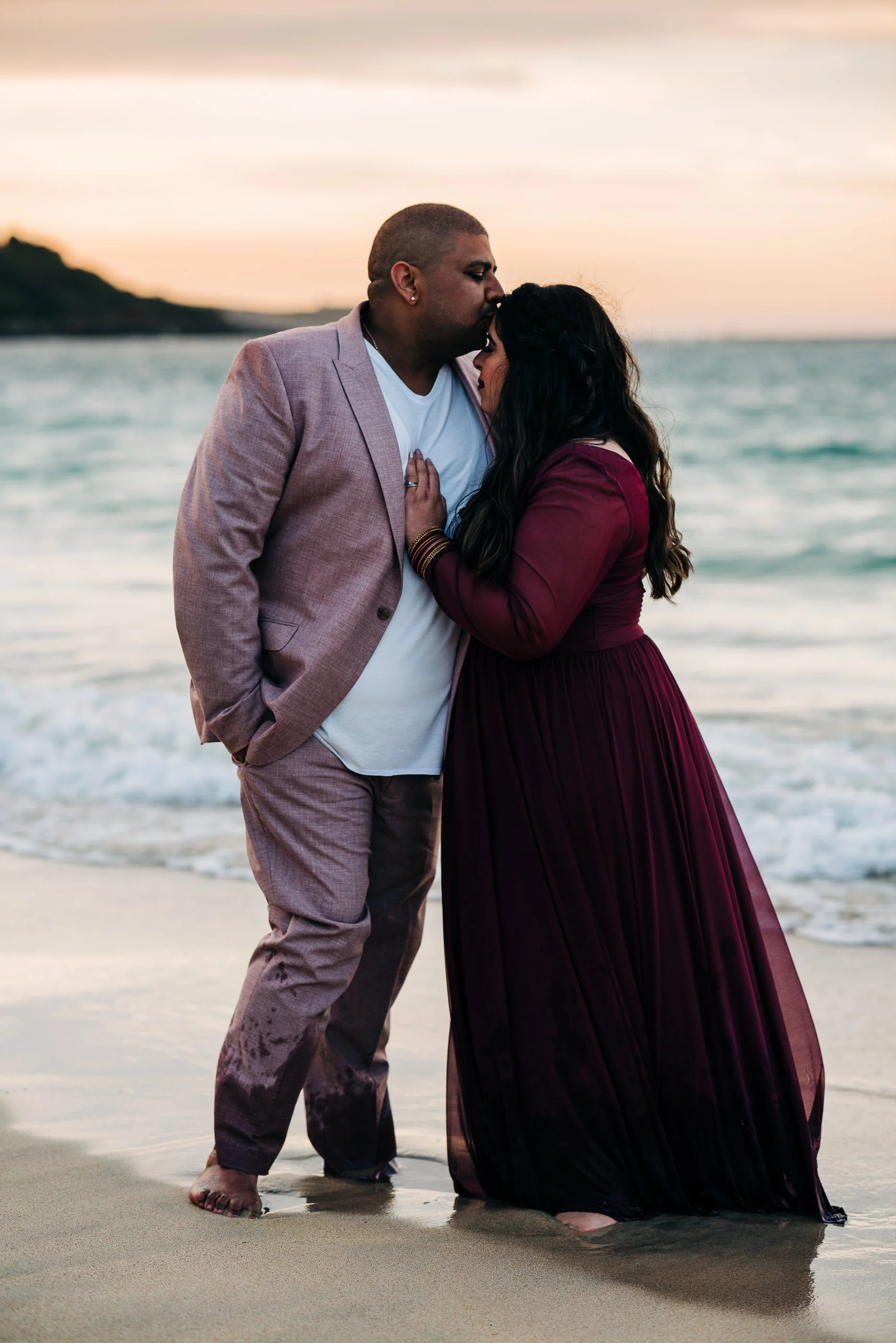 A couple standing on a beach at sunset, with the man kissing the woman's forehead. The man wears a light-colored suit, and the woman wears a long, flowing burgundy dress.