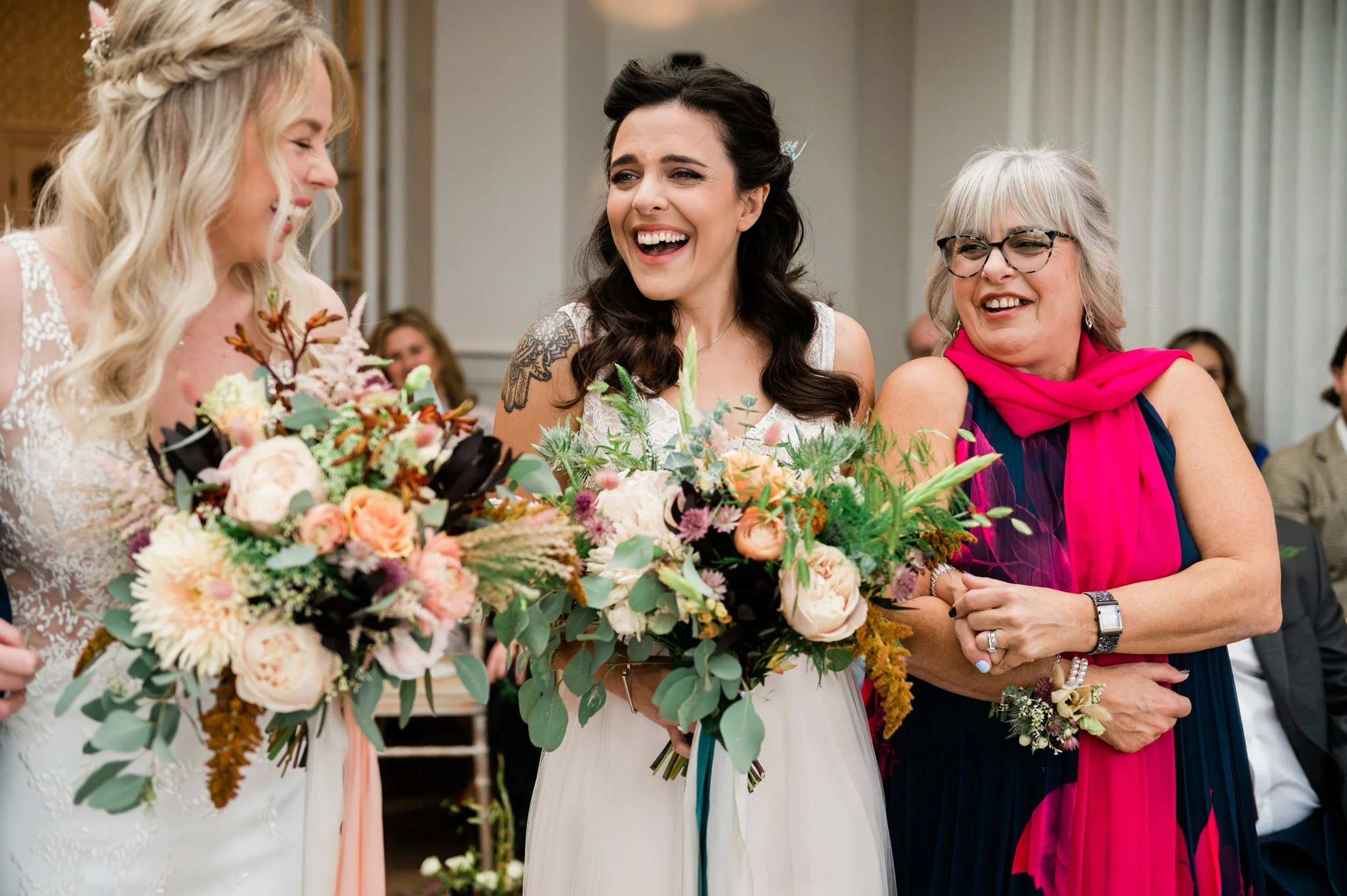 Three women at a wedding reception, two holding bouquets. One woman is in a white wedding dress, smiling and talking to the bride, who is in a white dress with dark hair. An older woman in a dark dress with a pink scarf around her neck smiles and hol