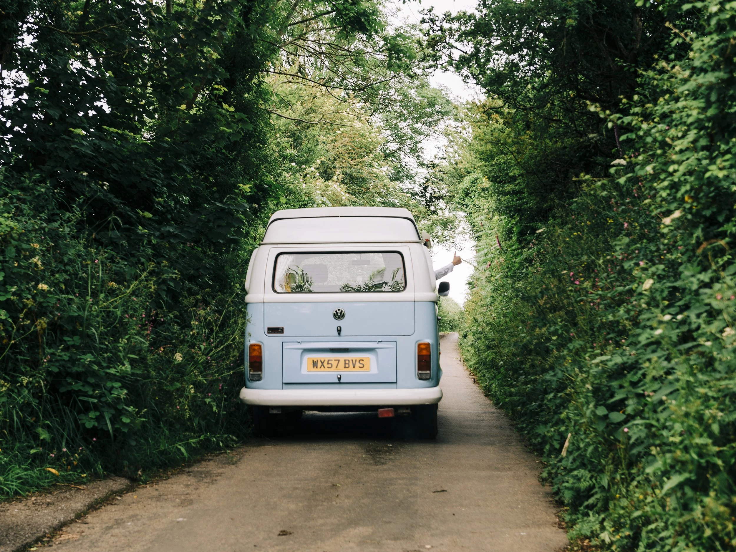 A light blue vintage Volkswagen van parked on a narrow dirt road surrounded by dense green foliage and trees, with a person standing at the rear of the van, pointing towards the sky.
