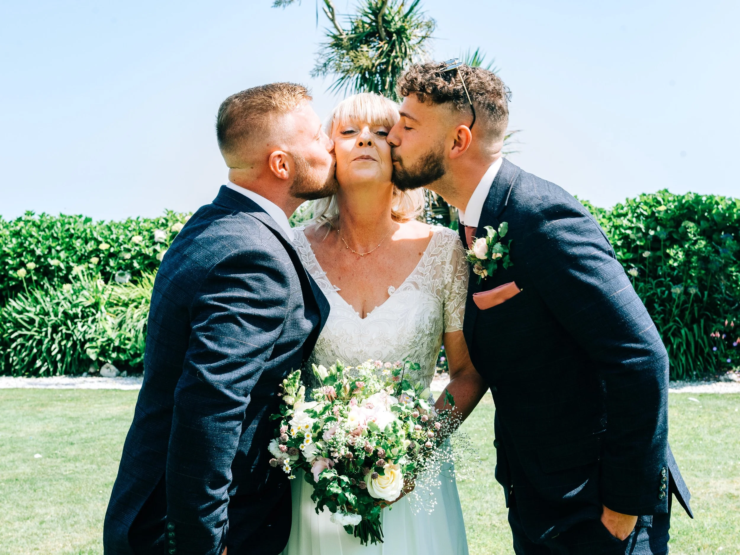 A woman in a wedding dress with blonde hair receiving kisses on each cheek from two men in suits, outdoors with greenery and palm trees in the background.