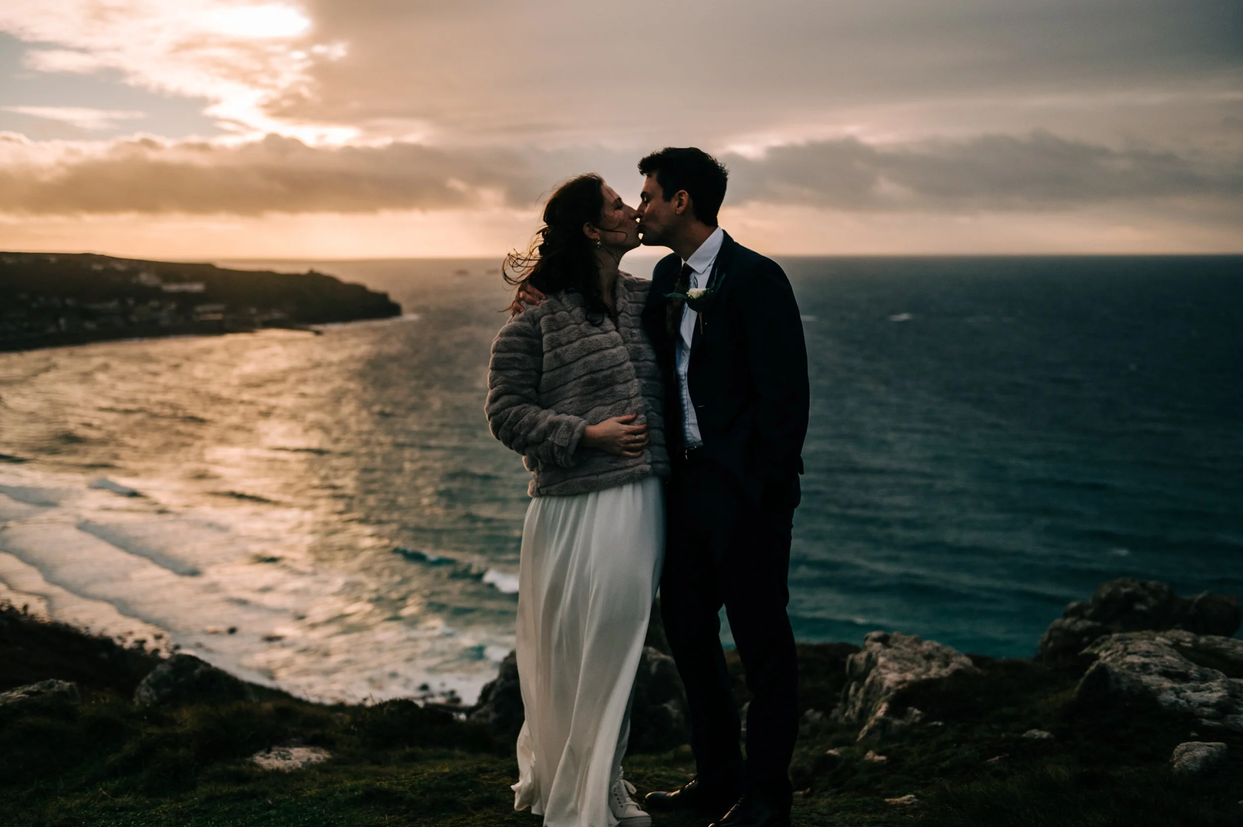 A pair of people, a woman in a white dress and a man in a suit, kissing near a sea cliff during sunset.