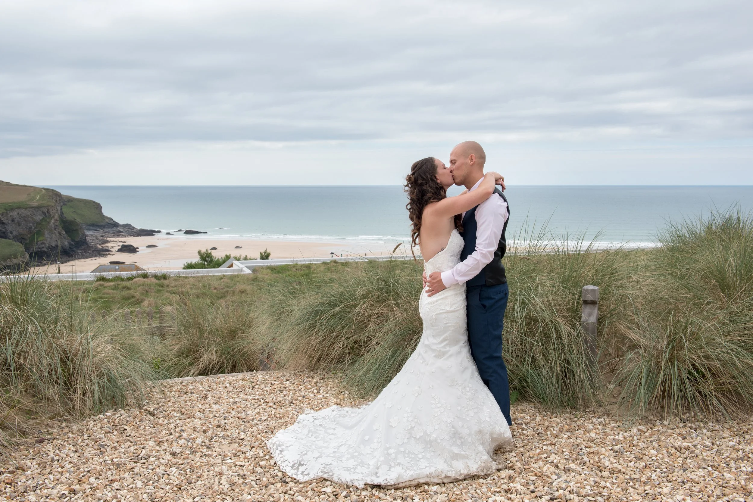 A bride and groom kissing on a gravel path near a beach with green dunes and cloudy sky in the background.