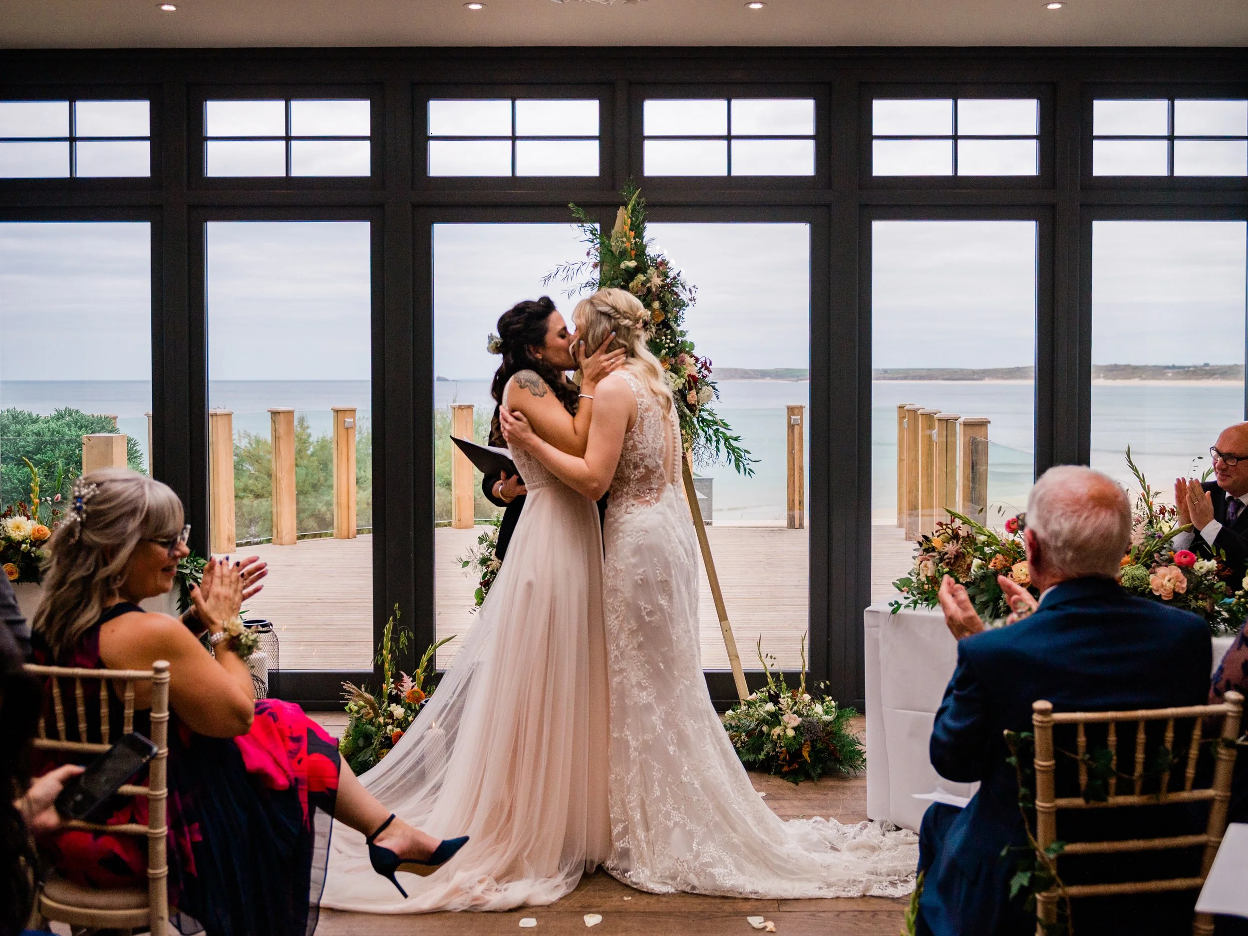 Two women in wedding dresses kissing during a wedding ceremony by a large window overlooking the ocean. Guests are seated on either side, clapping and smiling.