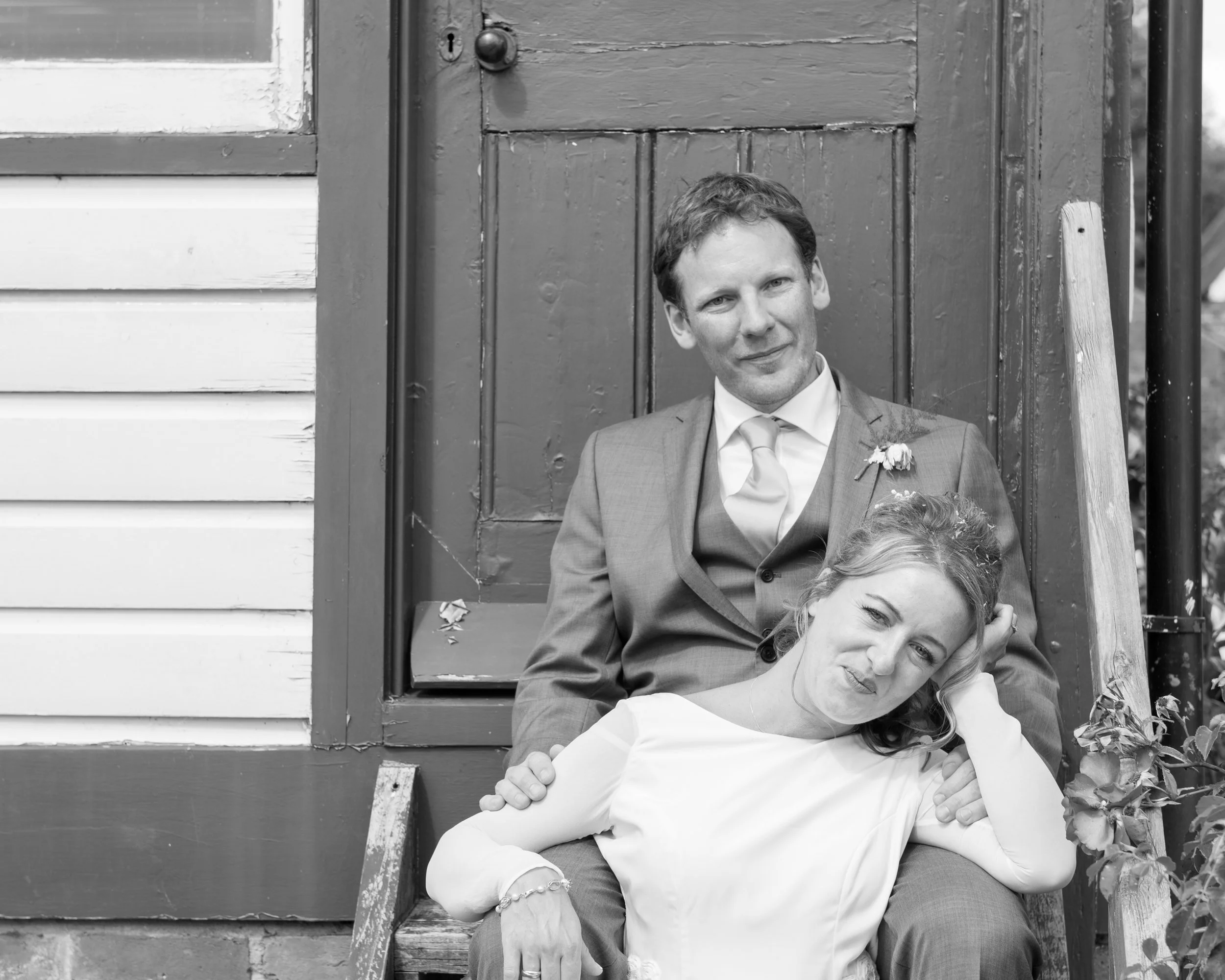 Black and white photo of a man in a suit sitting on stairs with a woman in a white dress leaning against him, both smiling and looking at the camera.