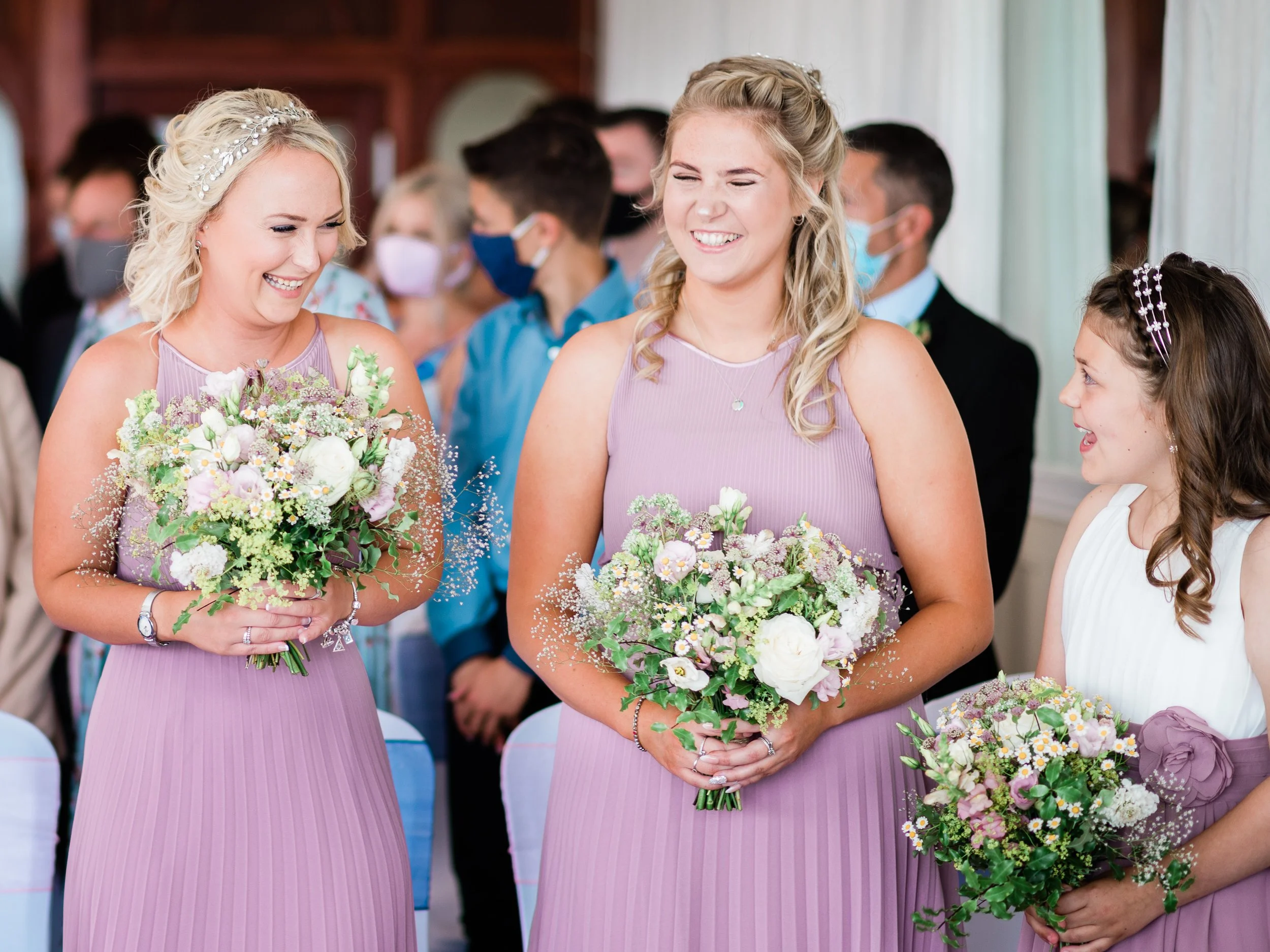 Three women, wearing lavender dresses, smiling and holding bouquets at a wedding celebration